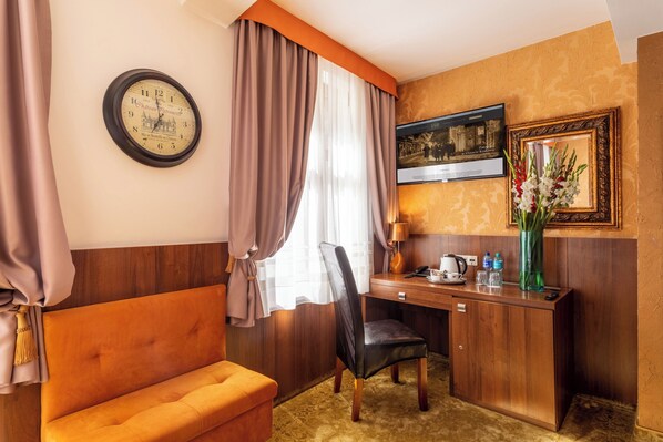 Une chambre d’hôtel avec un bureau en bois, une chaise, un canapé, une horloge, un tableau encadré et un vase avec des fleurs.