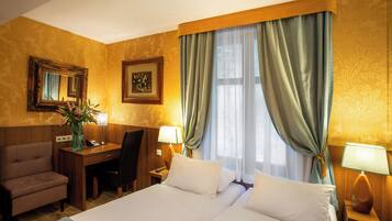 A hotel room with a large bed, a desk with a chair, a small table with a lamp, and a mirror on the wall.
