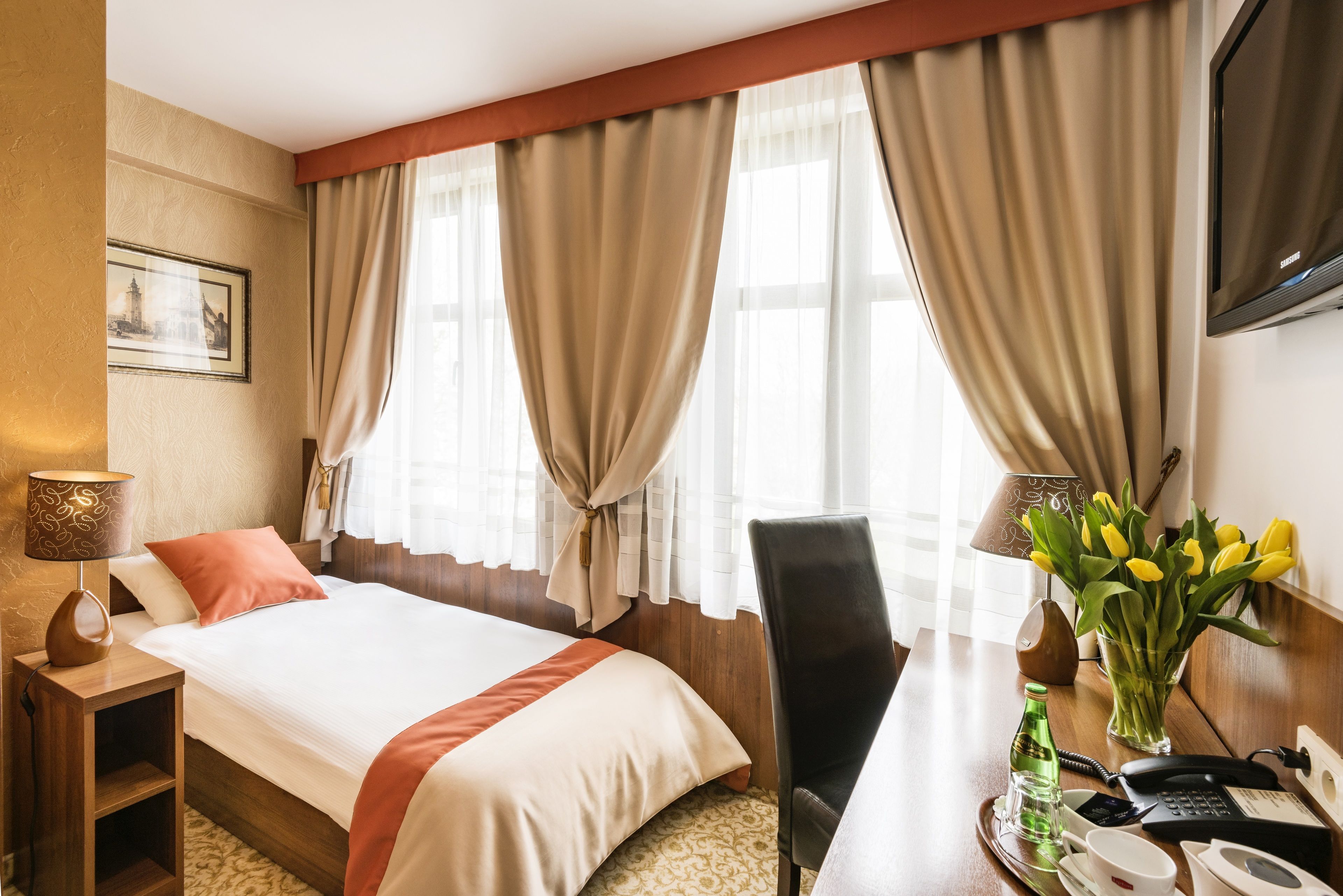 A hotel room with a large bed, a desk with a telephone and a vase of flowers, and a television on the wall.