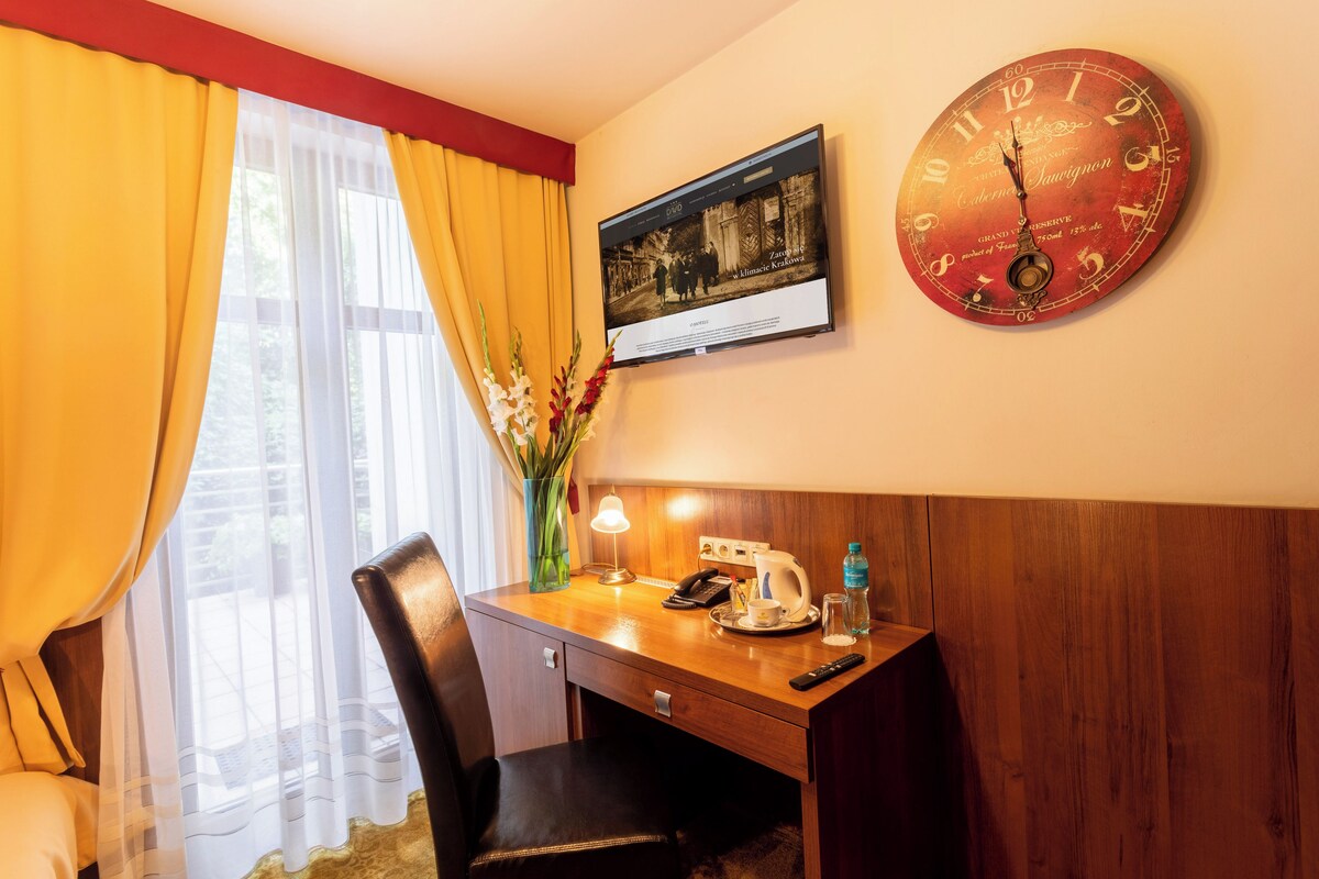 A hotel room with a wooden desk, a clock, a framed picture, and a window with curtains.