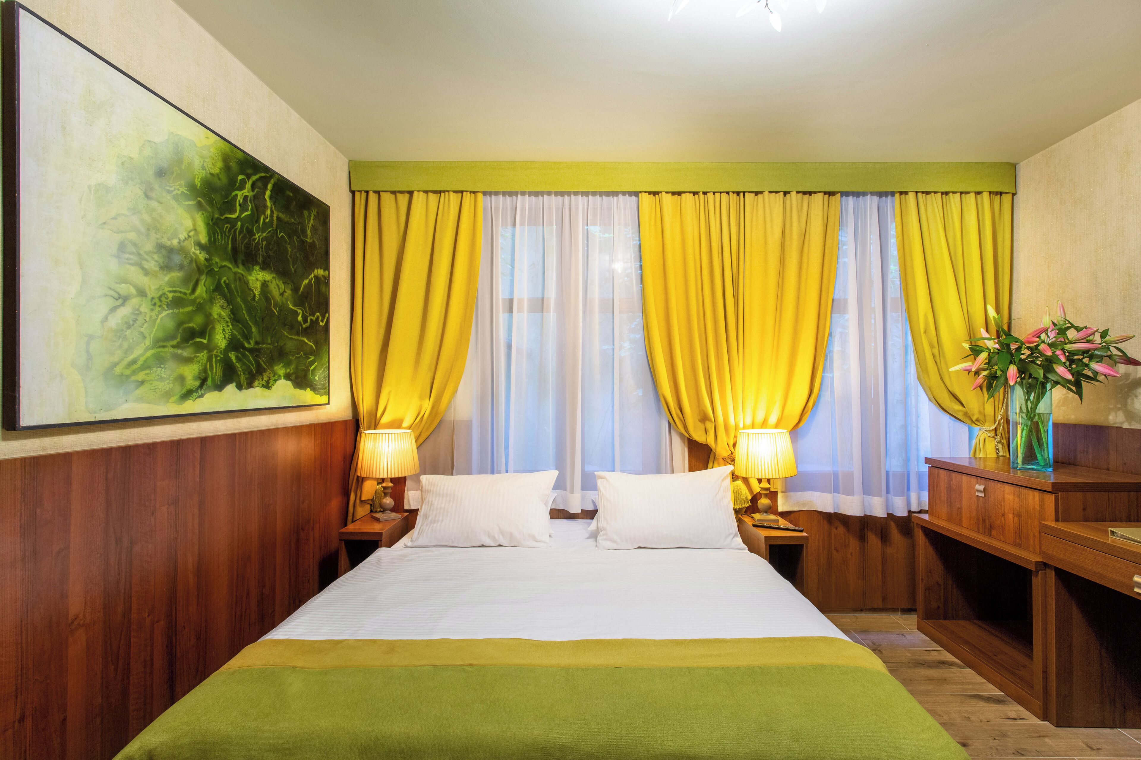 A hotel room with a large bed, two bedside tables with lamps, a wooden dresser, a painting on the wall, and yellow curtains.
