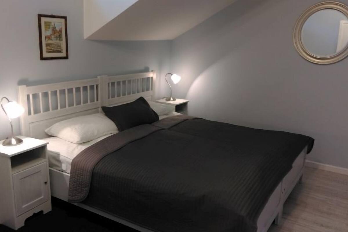 A bedroom with a white bed, two bedside tables with lamps, a framed picture on the wall, and a round mirror.