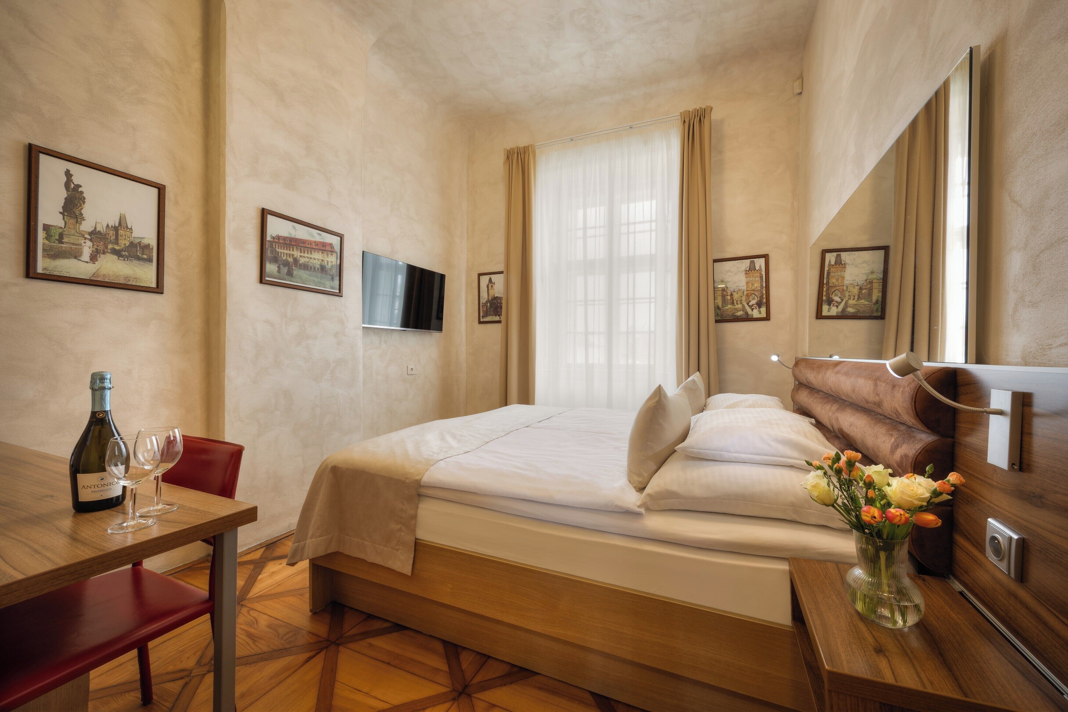 Ett hotellrum med en säng, ett skrivbord med en flaska och glas, en vas med blommor och inramade bilder på väggarna.