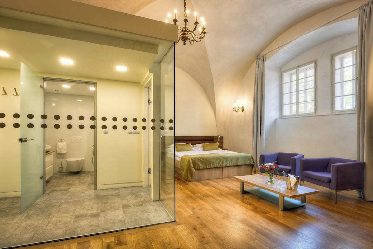 Quarto de hotel moderno com divisória de vidro, cama, sofá, mesa de centro e lustre.