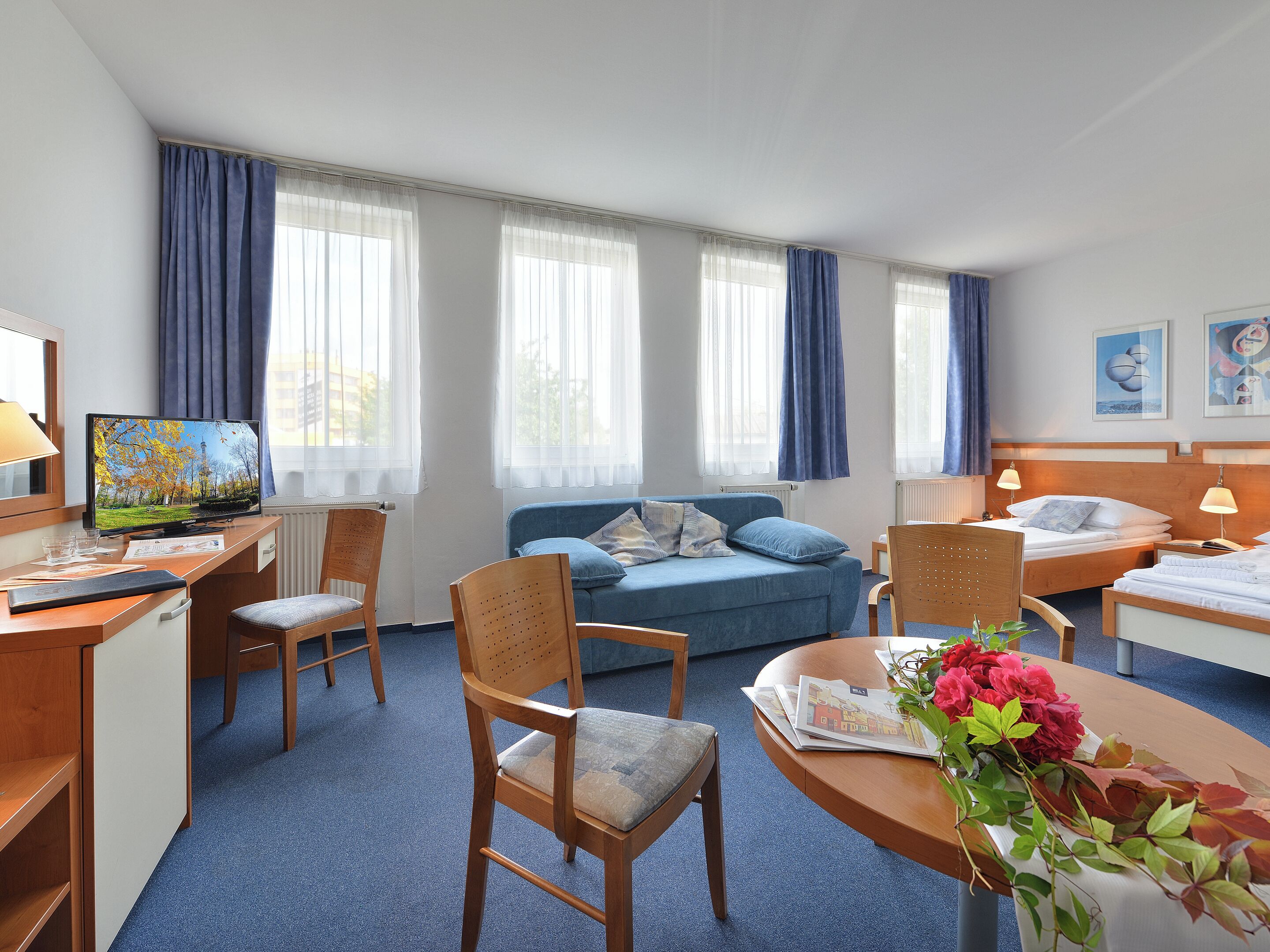 Quarto de hotel com escrivaninha, sofá azul, cama e televisão.