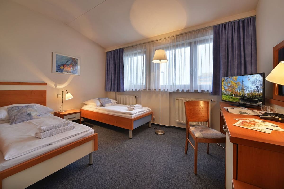 Quarto de hotel com duas camas, uma escrivaninha com cadeira, televisão e um quadro na parede.