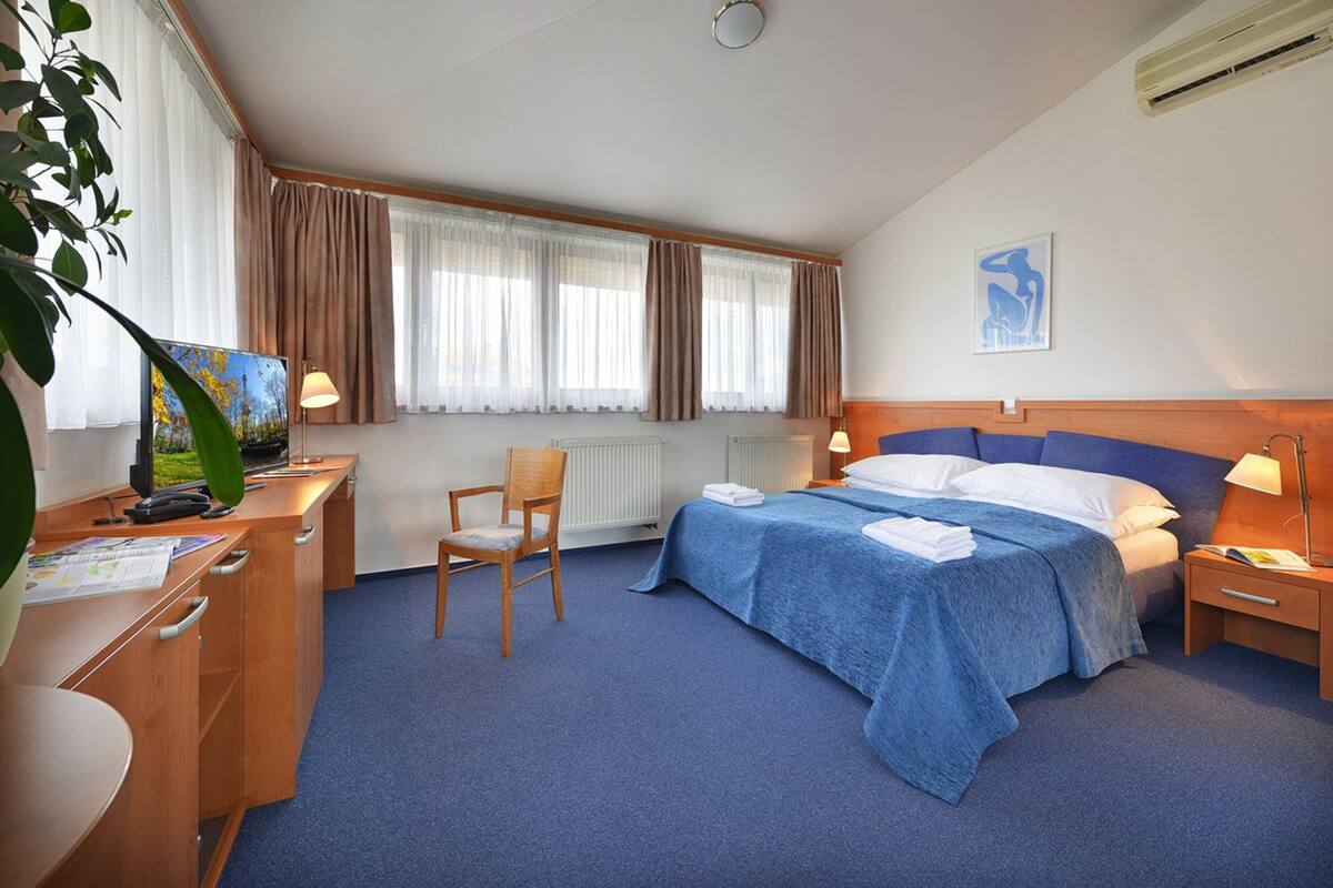 Quarto de hotel com cama, escrivaninha com computador, cadeira e janela com cortinas.
