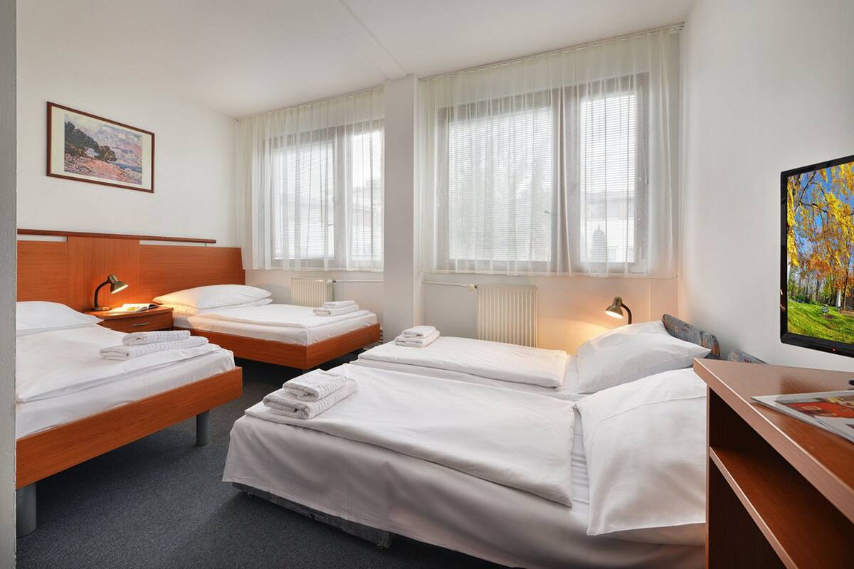 Quarto de hotel com três camas simples, cabeceira de madeira, escrivaninha com computador e televisão na parede.
