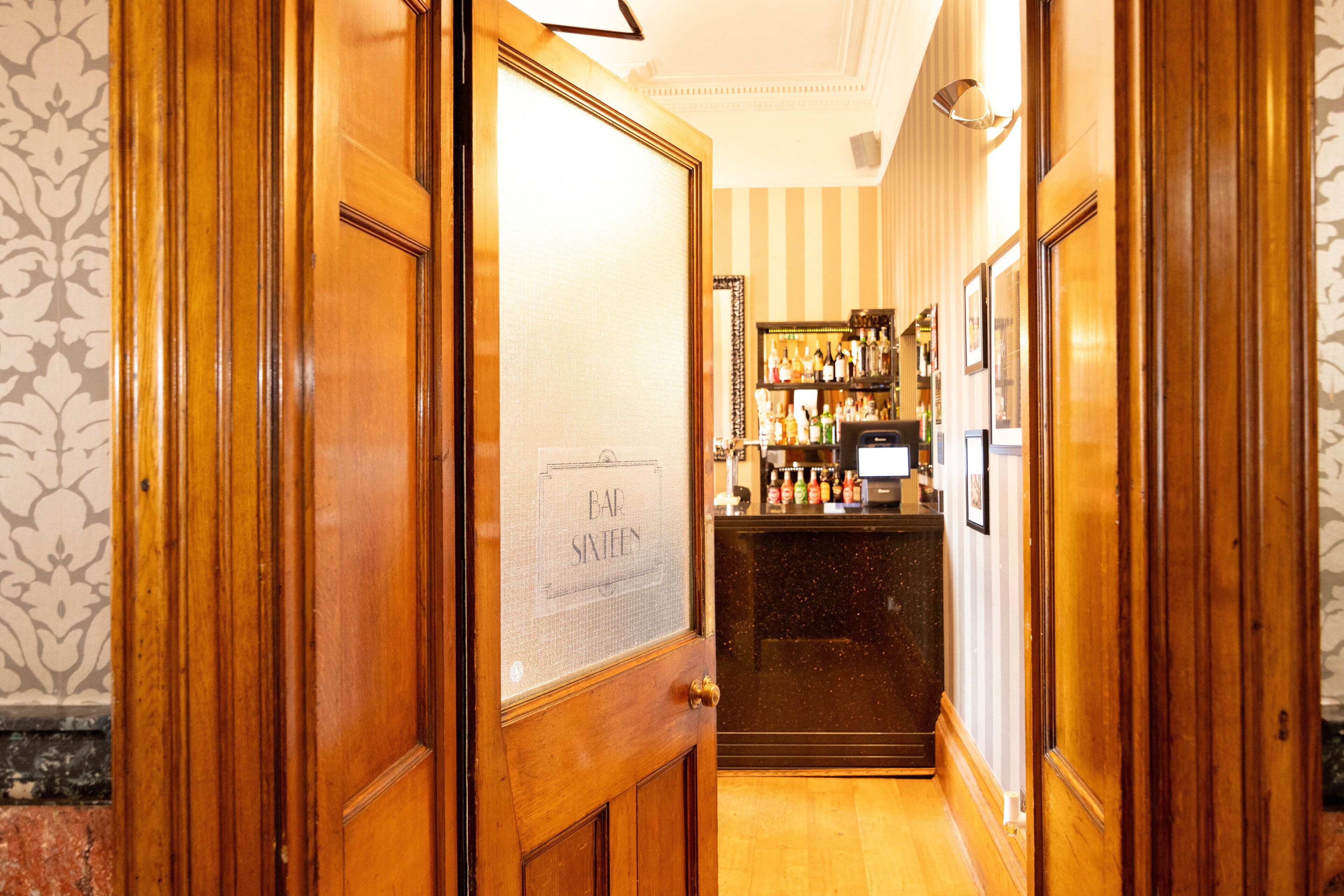 Un couloir avec des portes en bois, un coin bar avec des étagères remplies de bouteilles, et une enseigne indiquant « BAR SIXTEEN ».
