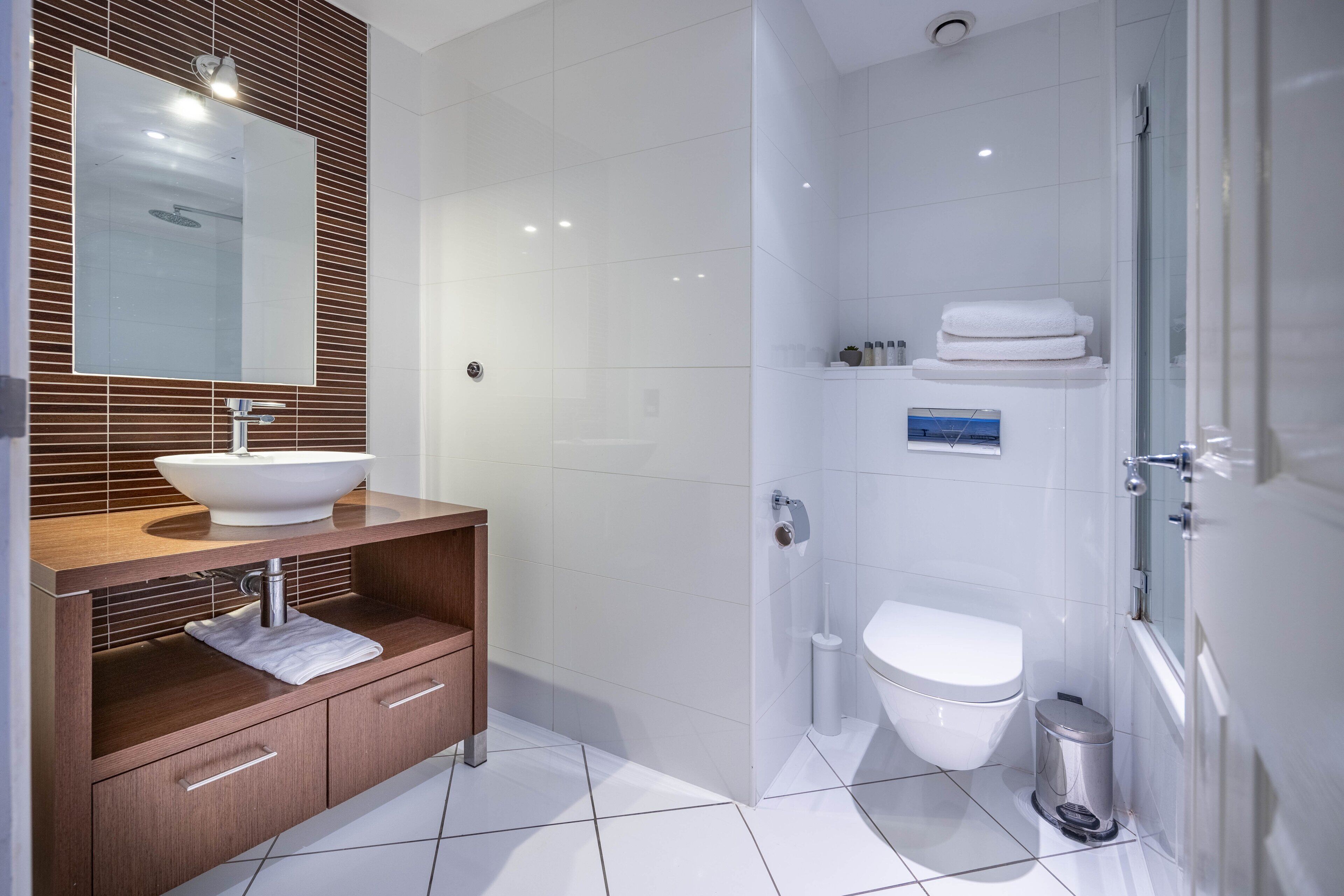 A modern bathroom with a wooden vanity, a large mirror, a white sink, a toilet, and a shower area.