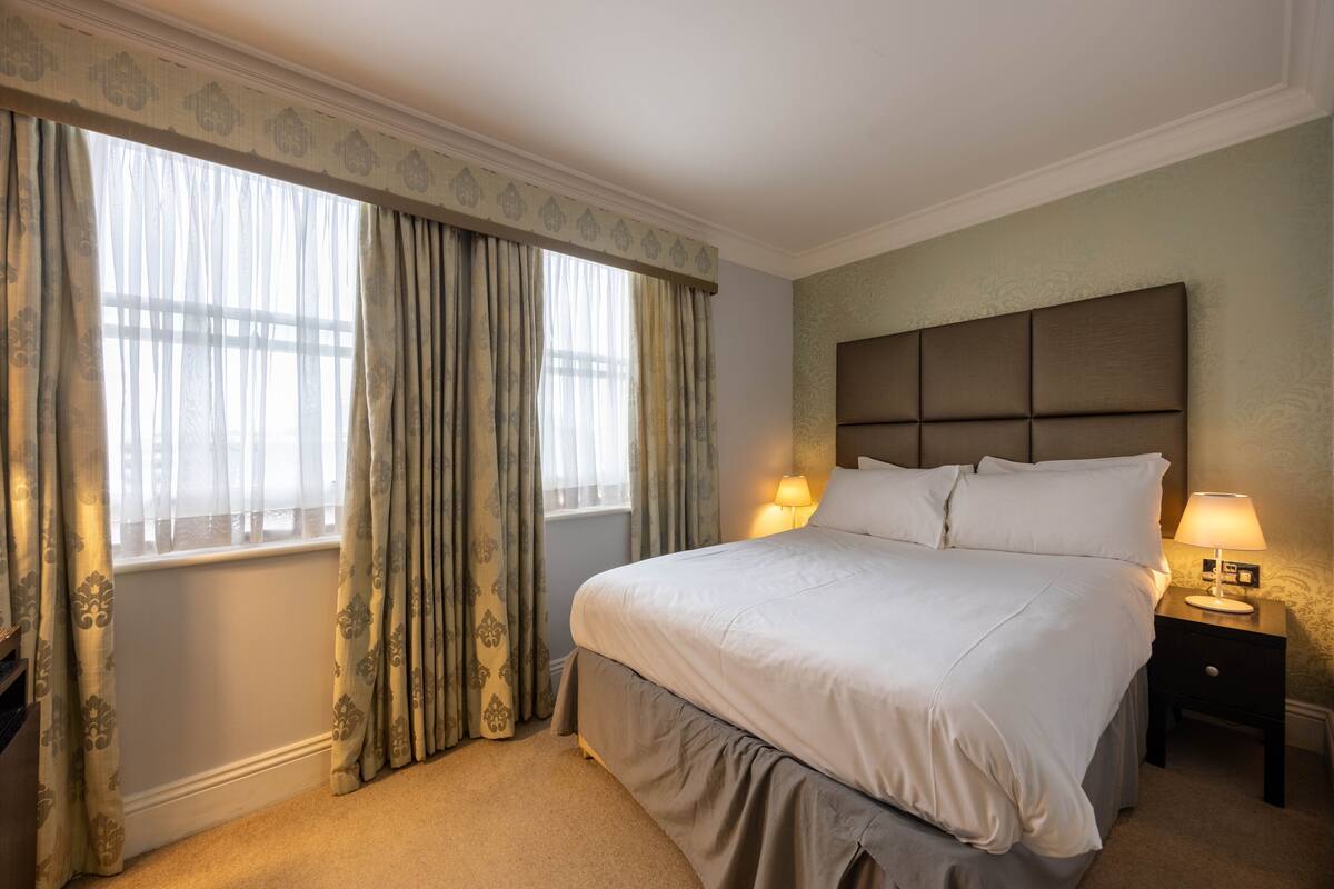 A hotel room with a large bed, a nightstand with a lamp, and a window with curtains.