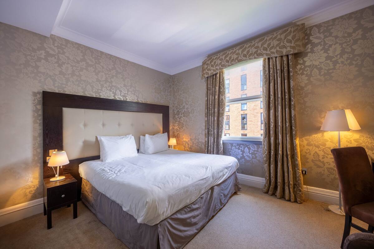 A hotel room with a large bed, a nightstand with a lamp, a chair, and a window with curtains.