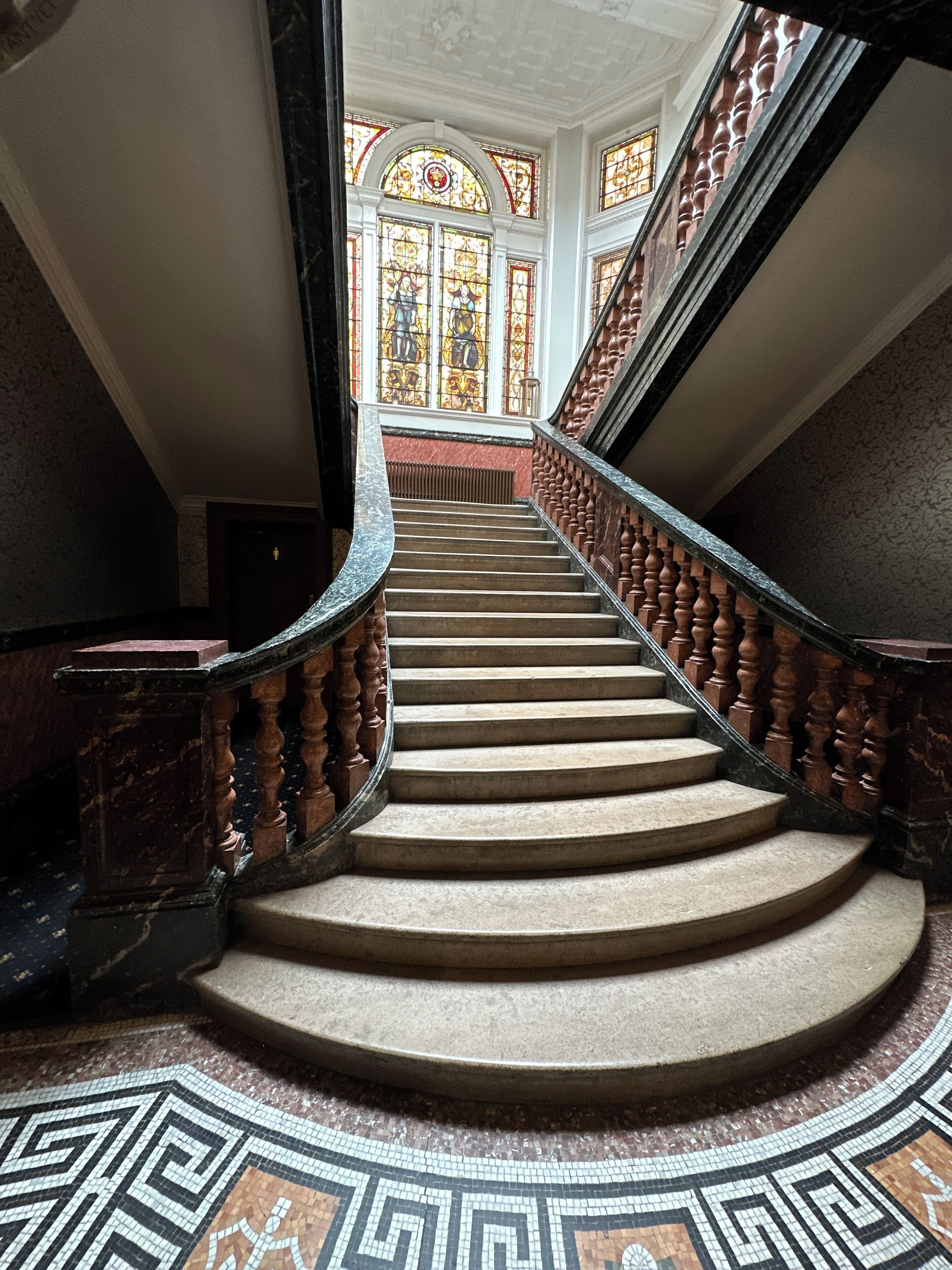 Eine imposante Treppe mit aufwendiger Balustrade und einem gemusterten Boden, die zu einem Raum mit großen Buntglasfenstern führt.