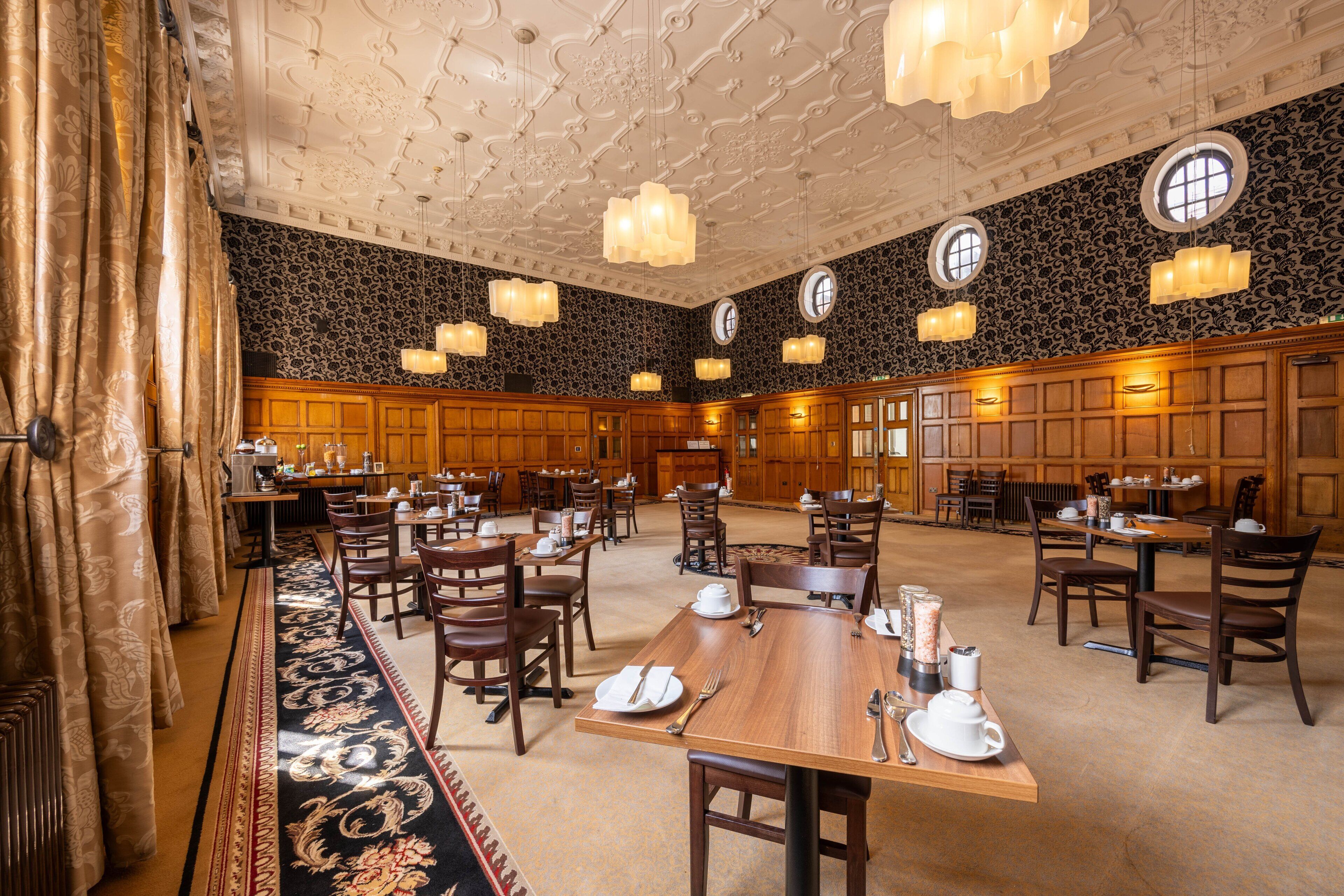Uma ampla sala de jantar com várias mesas e cadeiras, teto ricamente decorado e janelas grandes.