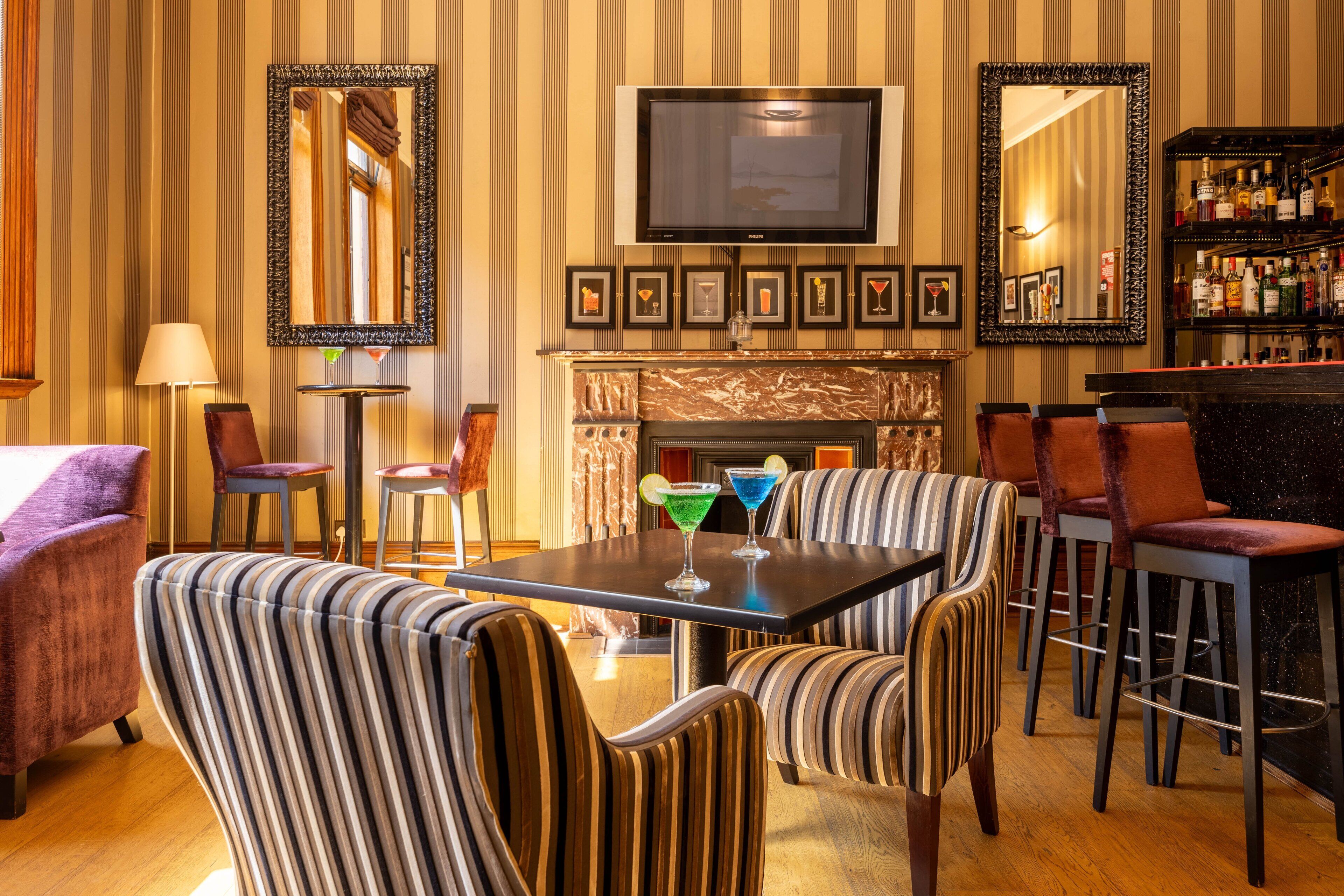 A bar area with striped upholstered chairs, a television above the fireplace, and framed pictures on the wall.