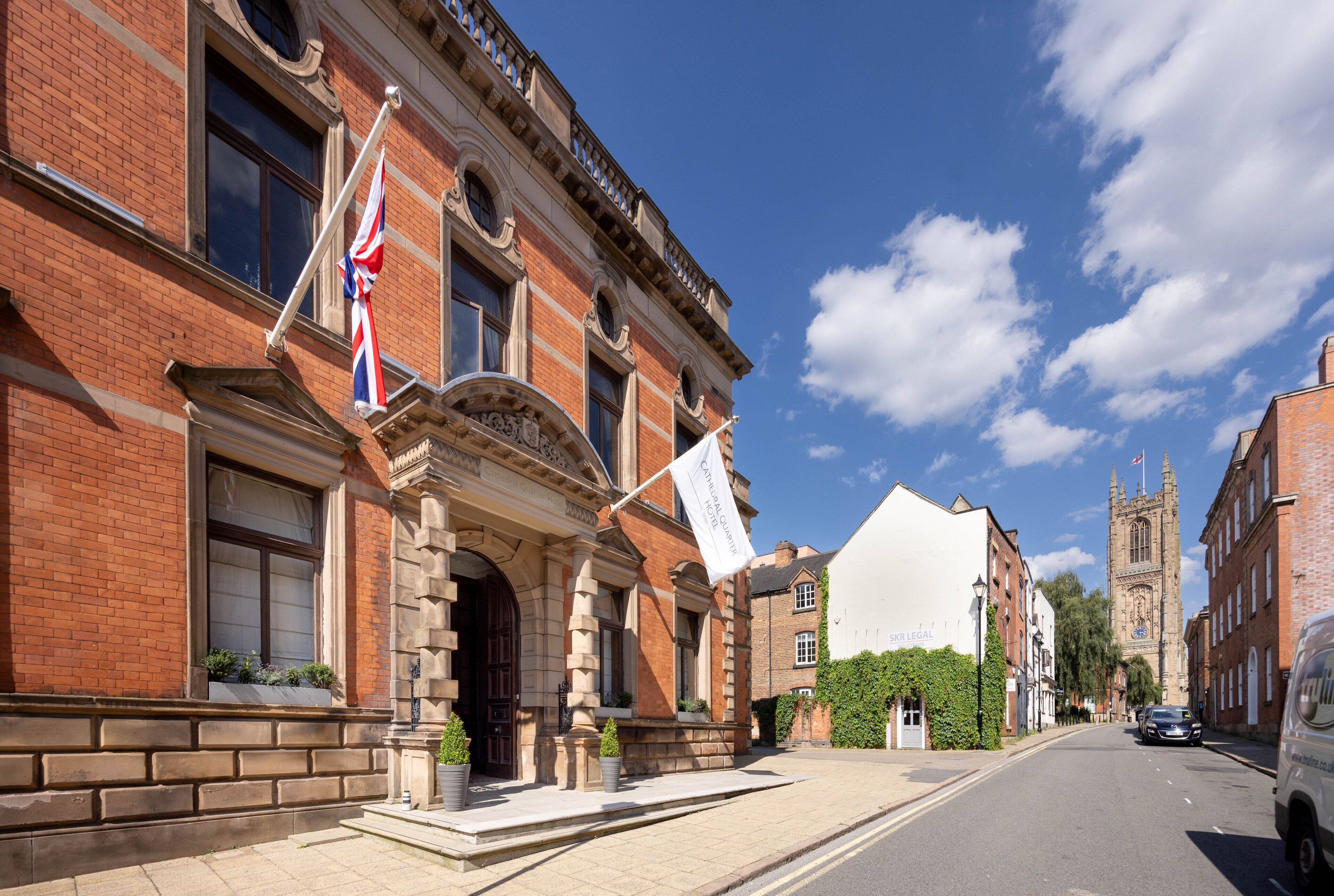 A historic building with a prominent entrance, a flag, and a church in the background.