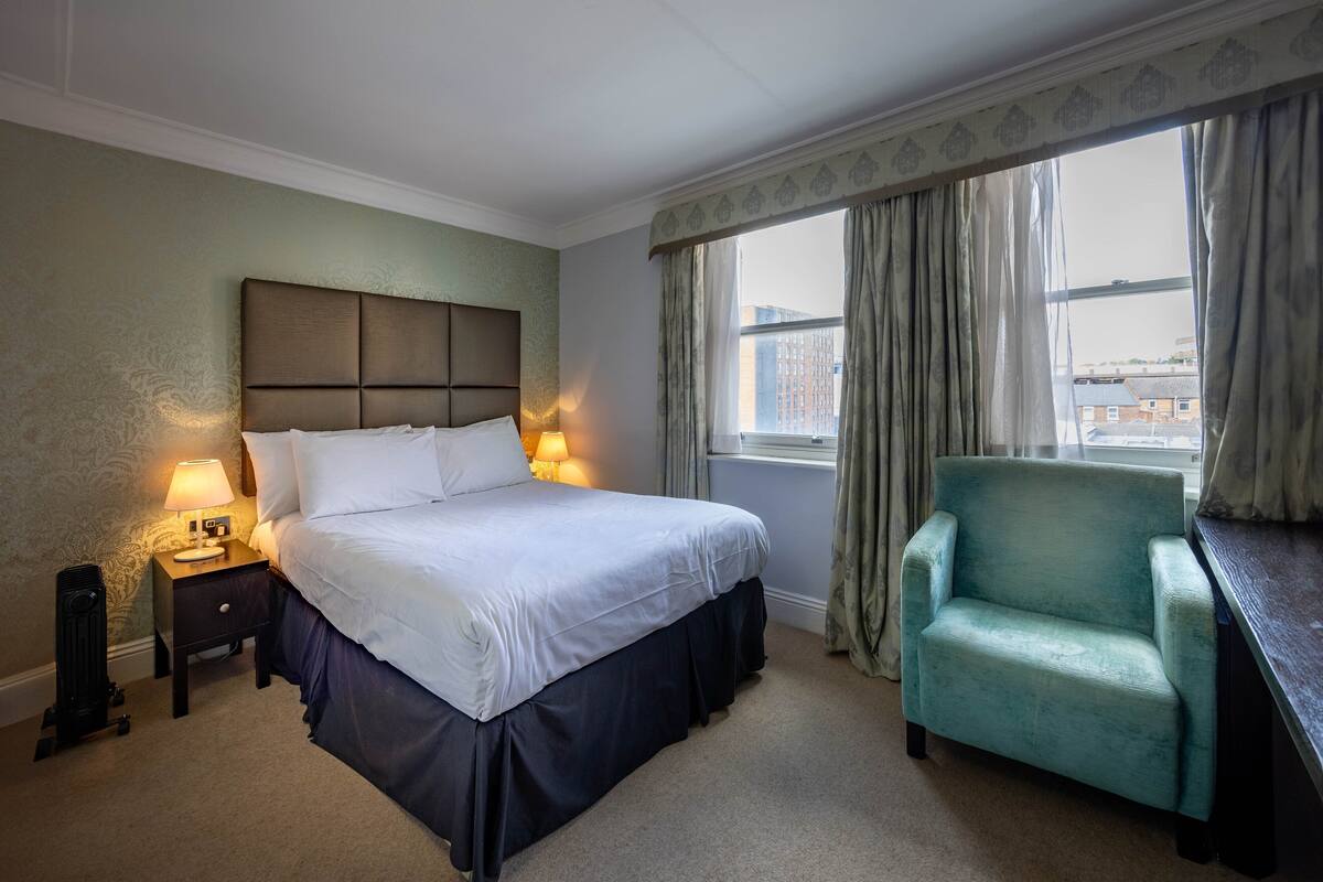 A hotel room with a large bed, a nightstand with a lamp, a chair, and a view of the city through the window.
