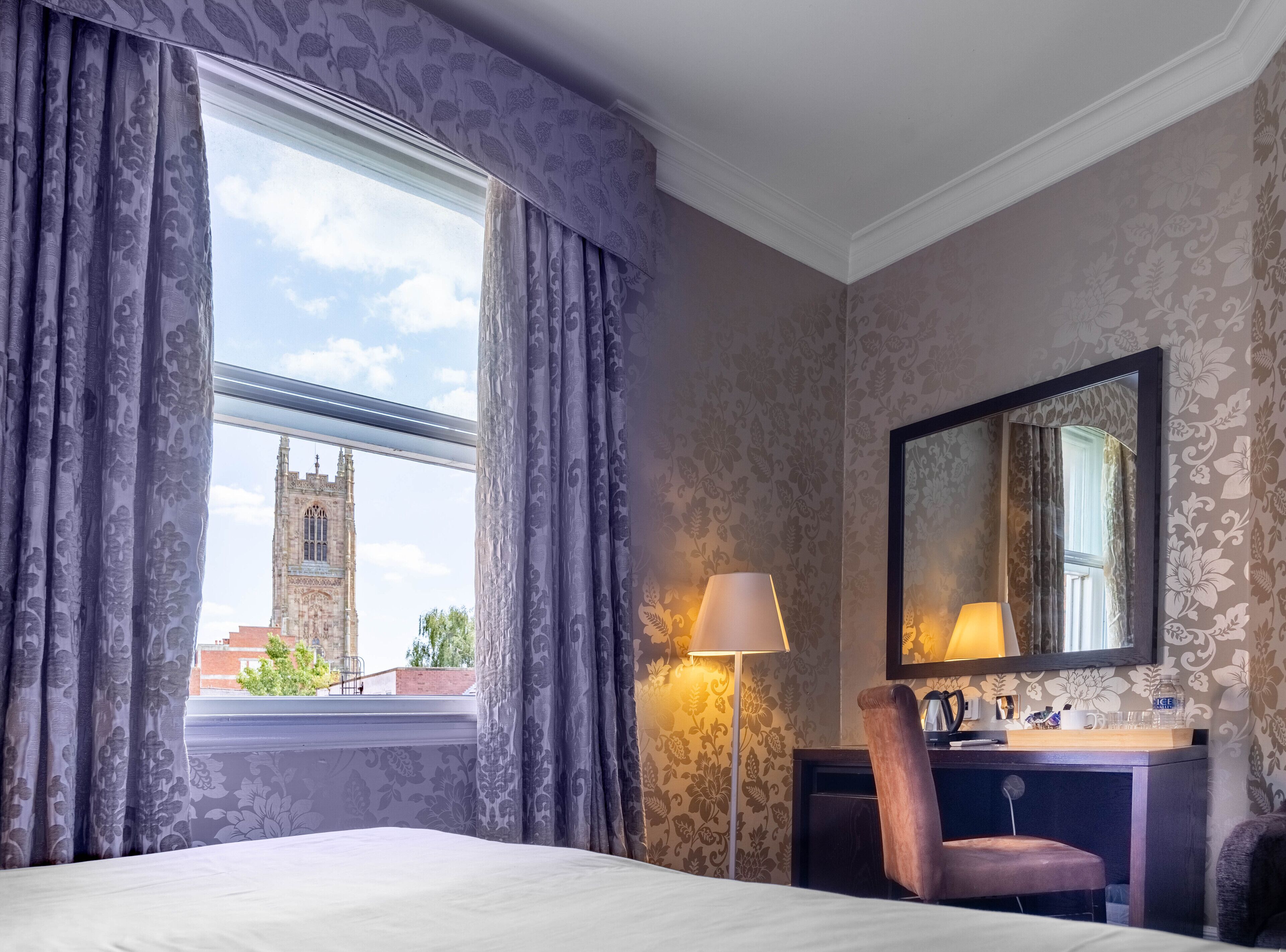 A hotel room with a large window, a desk with a lamp, a chair, and a mirror.