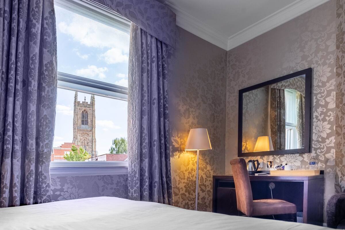 A hotel room with a large window, a desk with a lamp, a chair, and a mirror.