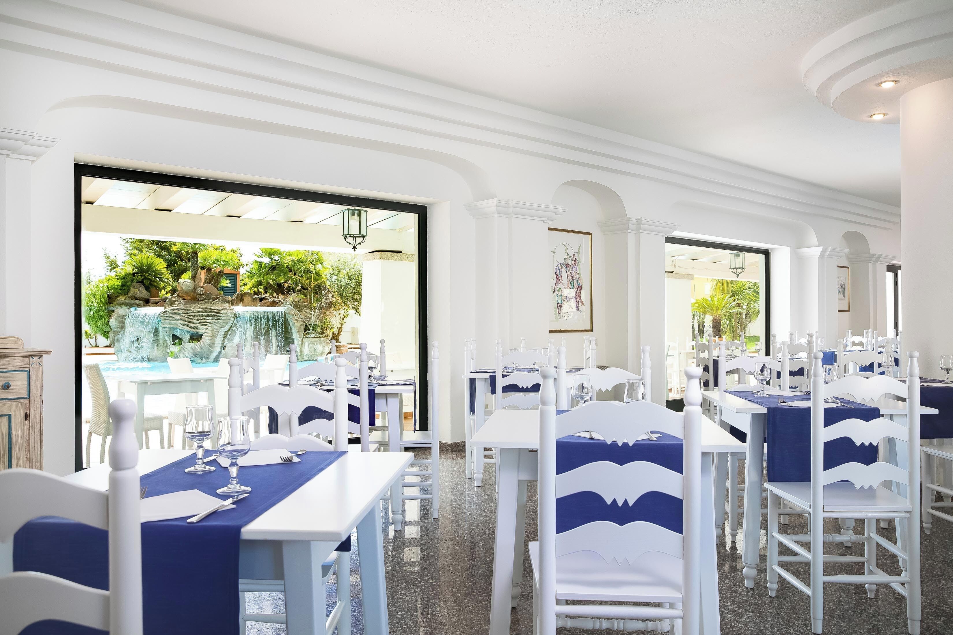 A restaurant with white tables and chairs, blue tablecloths, and a view of a garden through the open doors.