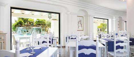 A restaurant with white tables and chairs, blue tablecloths, and a view of a garden through the open doors.