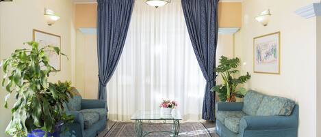 Una sala de estar con una mesa de centro de vidrio, dos sofás azules, una planta grande en maceta y una araña.
