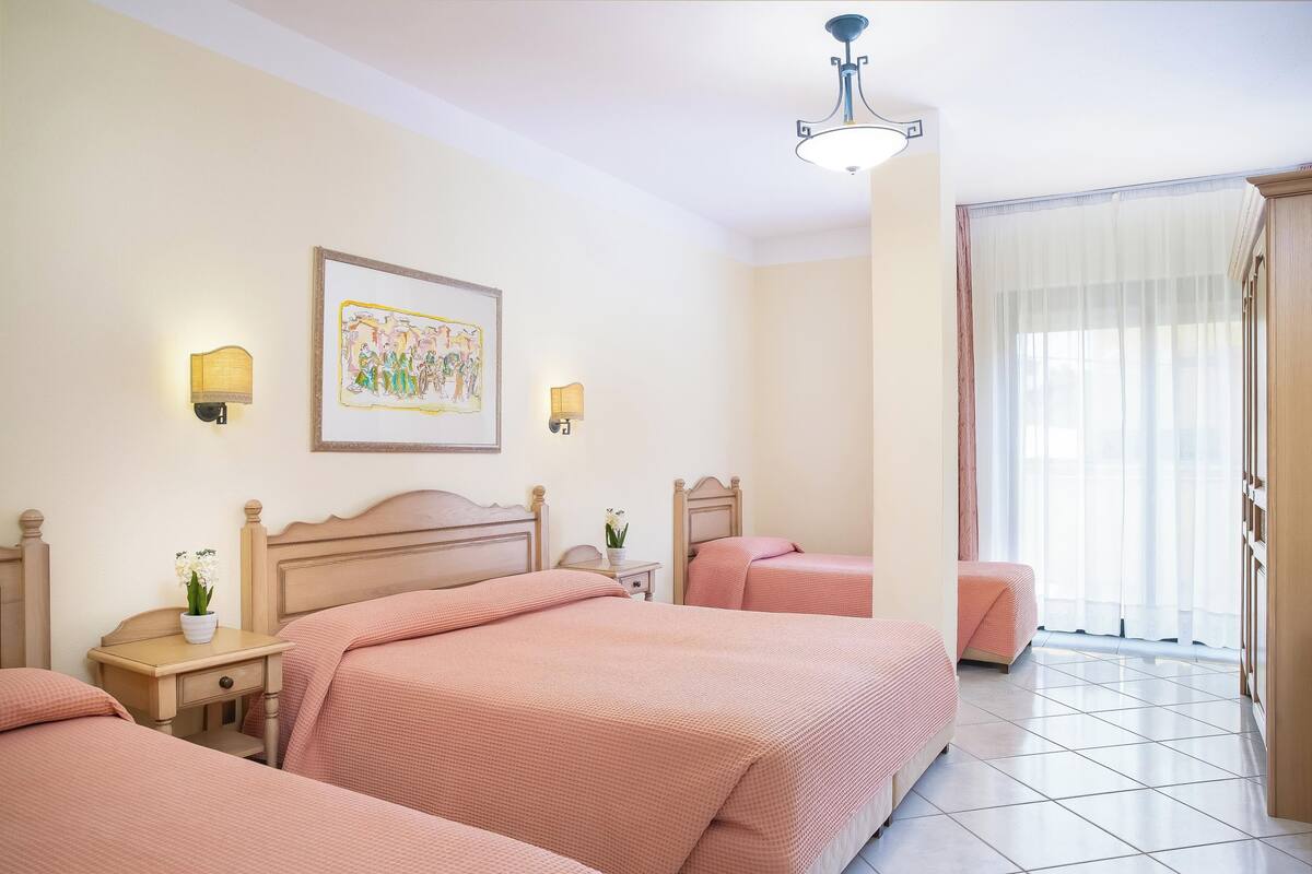 A hotel room with two beds, a nightstand with a lamp, a painting on the wall, and a large window with curtains.