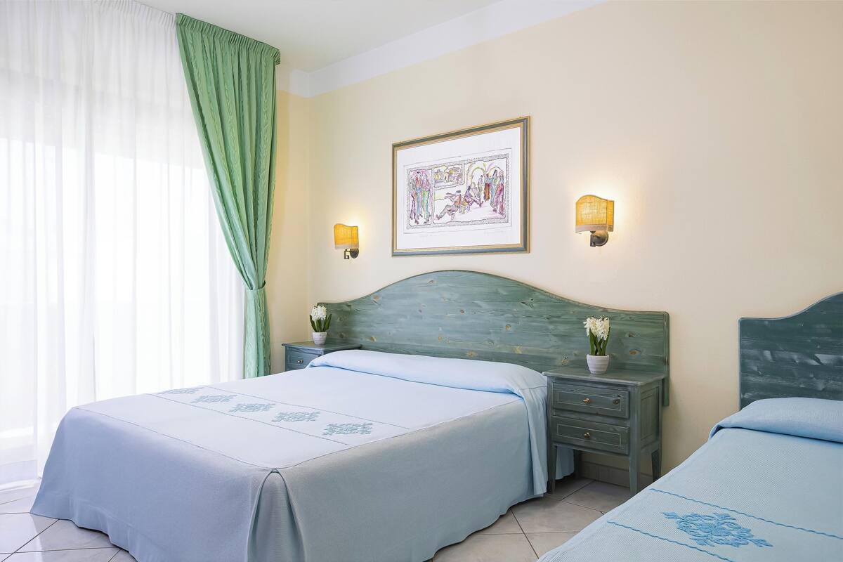 A hotel room with two beds, a green headboard, a framed picture on the wall, and two wall-mounted lamps.