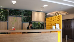 Una recepción con mostrador de madera, una recepcionista y un panel decorativo amarillo con diseños recortados.