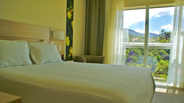 Habitación de hotel con una cama grande, una silla, una mesita de noche y una ventana con vistas a zonas verdes y montañas.