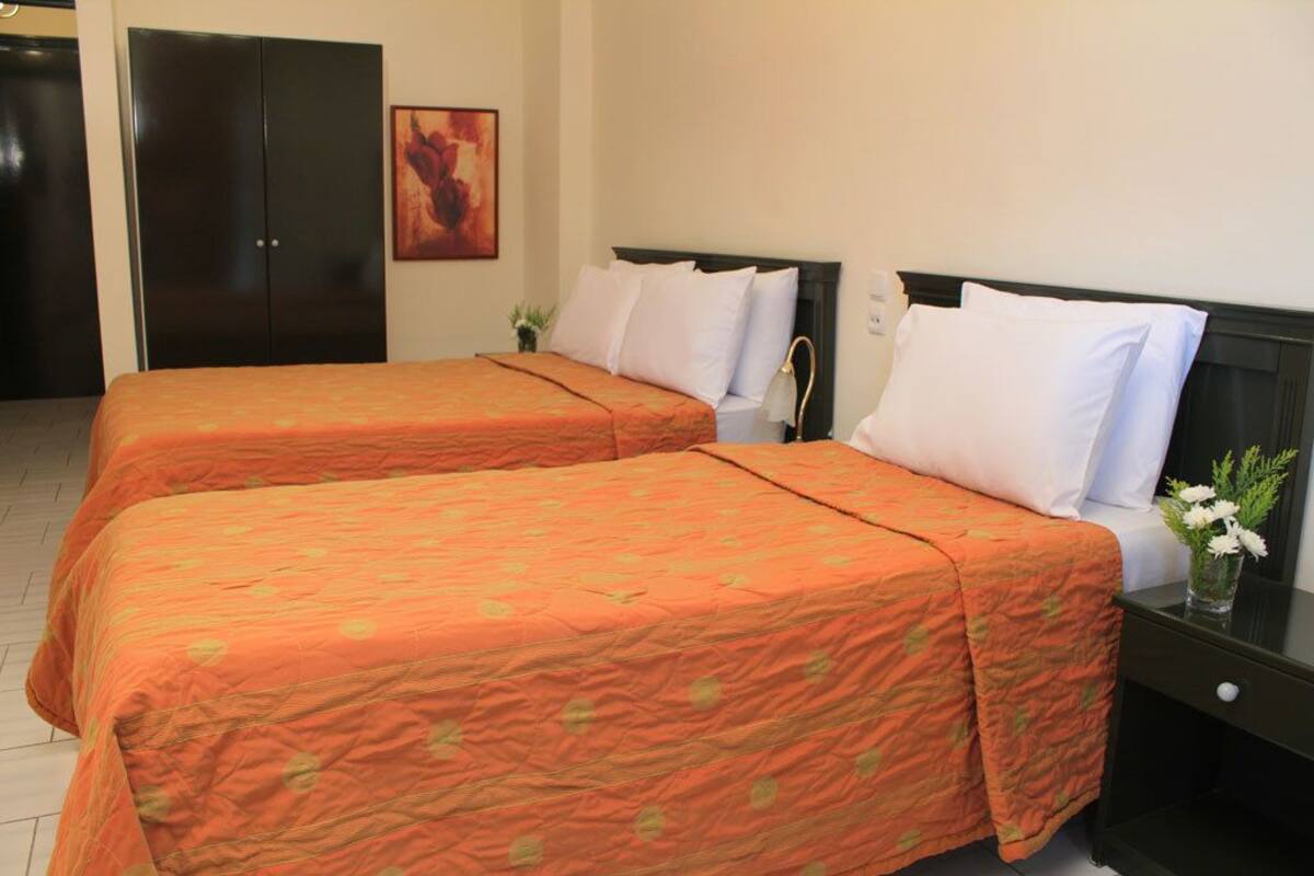 A hotel room with two beds, each with an orange bedspread and white pillows. There is a dark wooden wardrobe, a small table with a vase of flowers, and a painting on the wall.