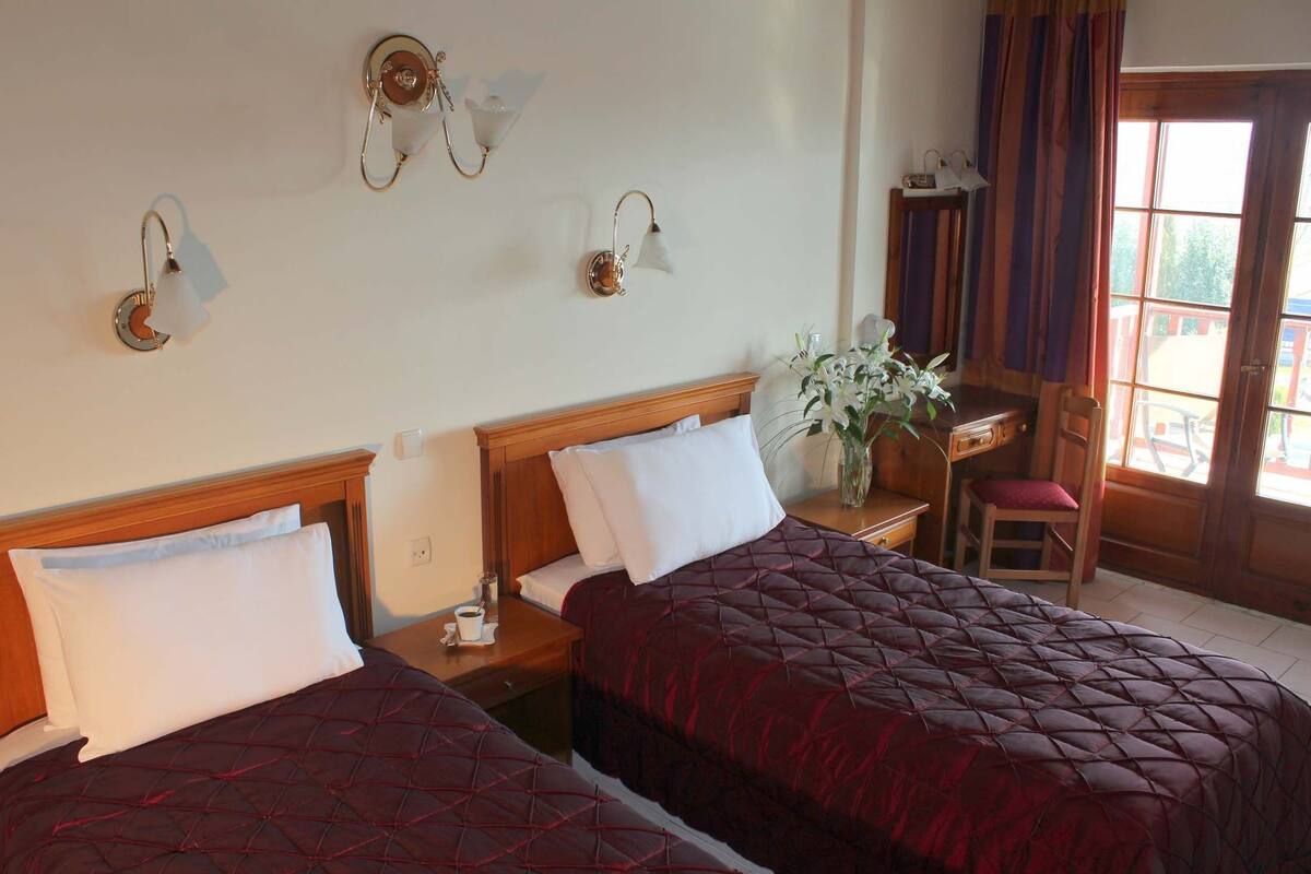 A hotel room with two beds, a wooden headboard, a nightstand with a lamp, a desk with a chair, and a window with curtains.