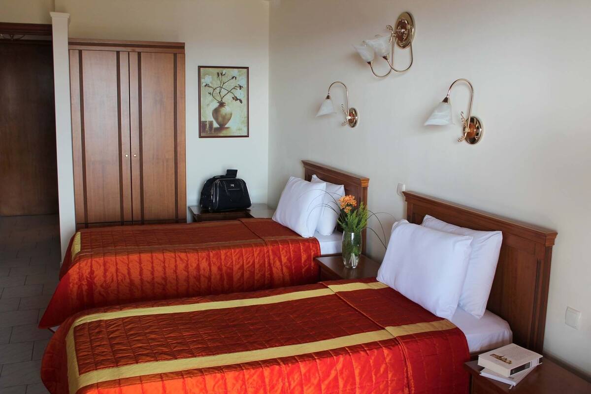 A hotel room with two beds, a wooden wardrobe, a desk with a lamp, and a painting on the wall.
