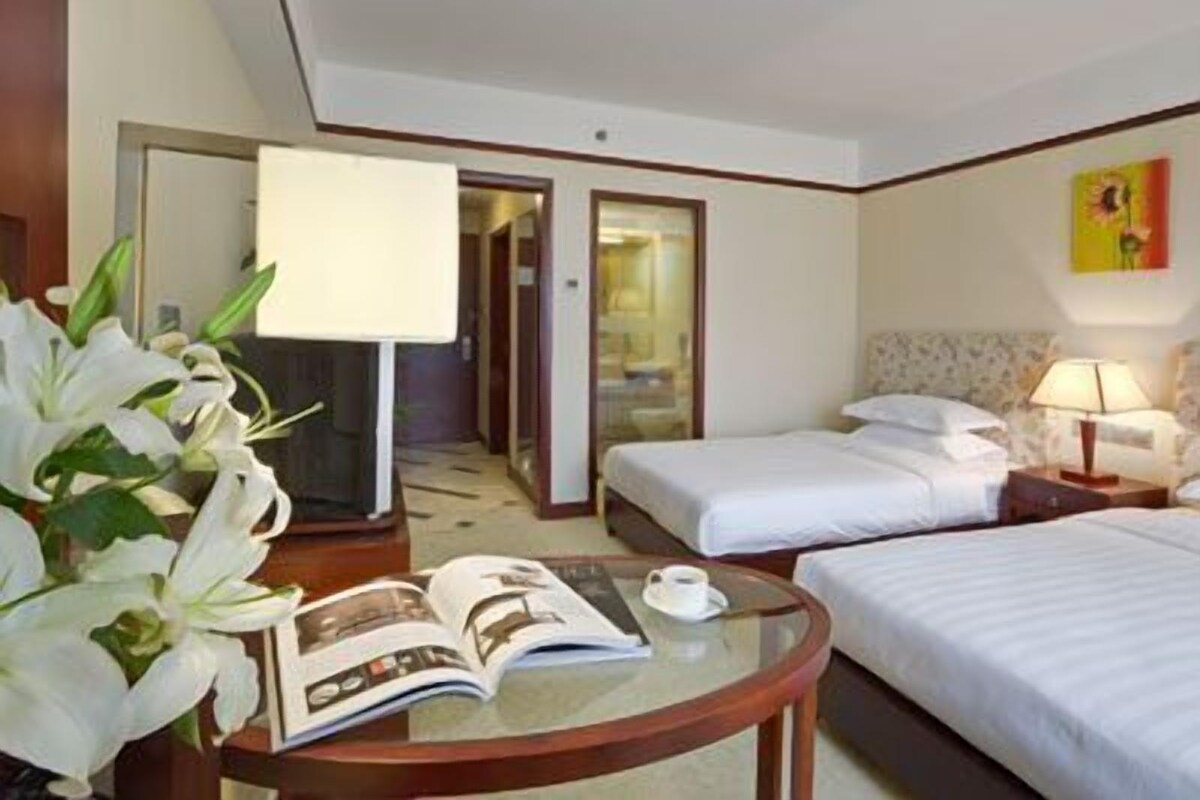 A hotel room with two beds, a glass coffee table with a magazine and a cup, a lamp, and a flower arrangement.