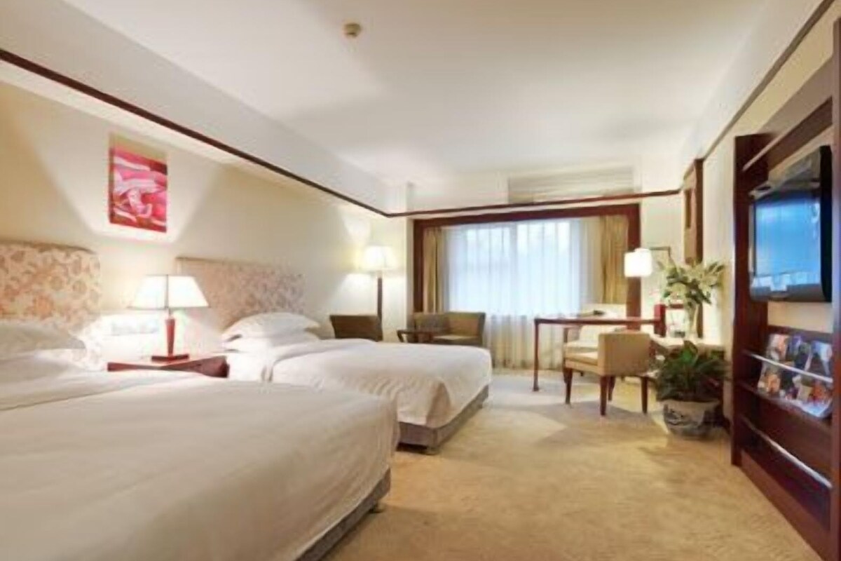 A hotel room with two beds, a desk, a chair, a TV, and a plant.