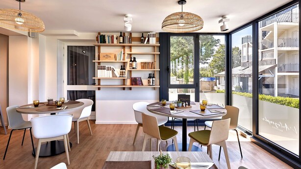A modern dining area with round tables, white chairs, and a large window offering a view of a building and greenery. - ibis La Roche sur Yon (Mouilleron-le-Captif)