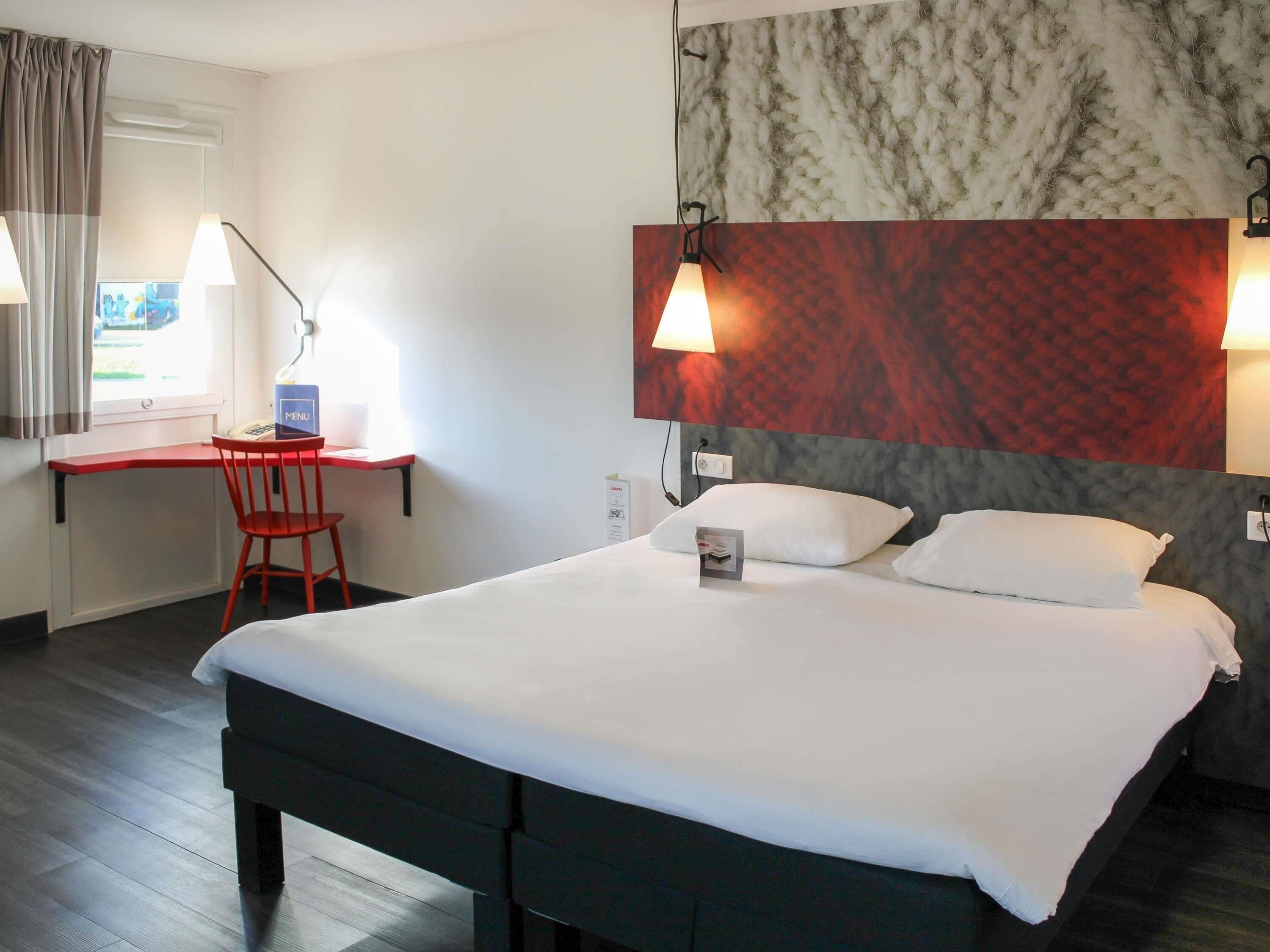 A hotel room with a bed, a desk with a chair, a window with curtains, and a wall with a textured pattern.