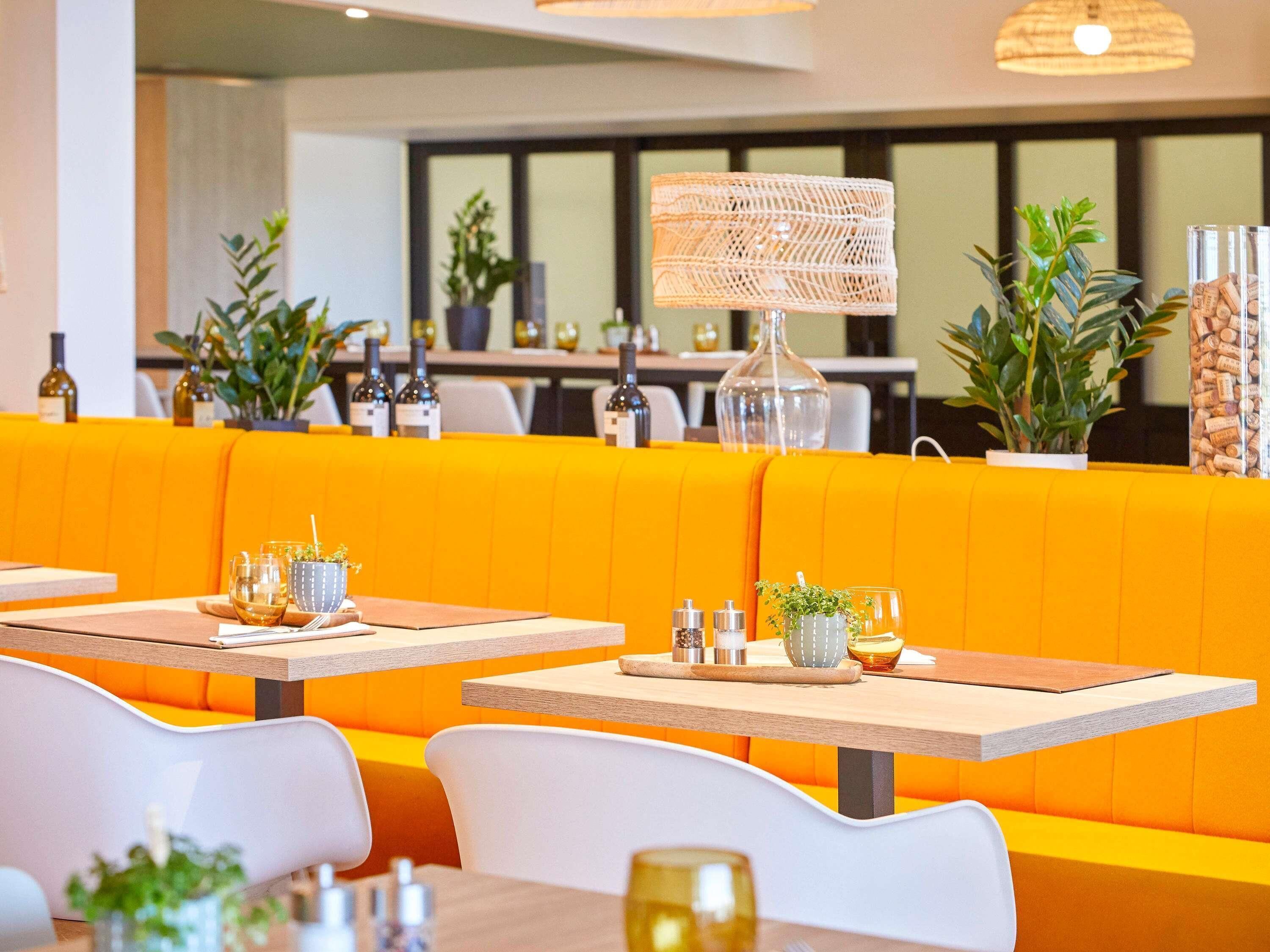 Un restaurante con asientos tipo banca color naranja y sillas blancas, mesas con copas de vino y botellas.