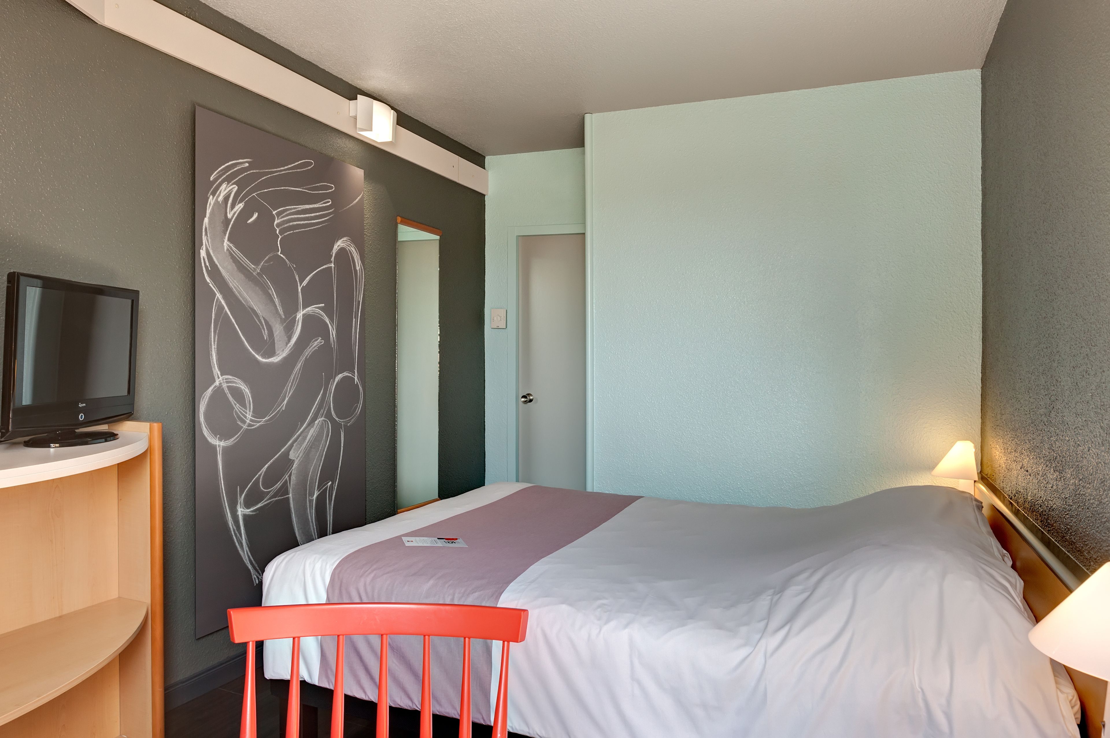 A hotel room with a bed, a desk with a television, a red chair, and a wall with a chalkboard drawing.