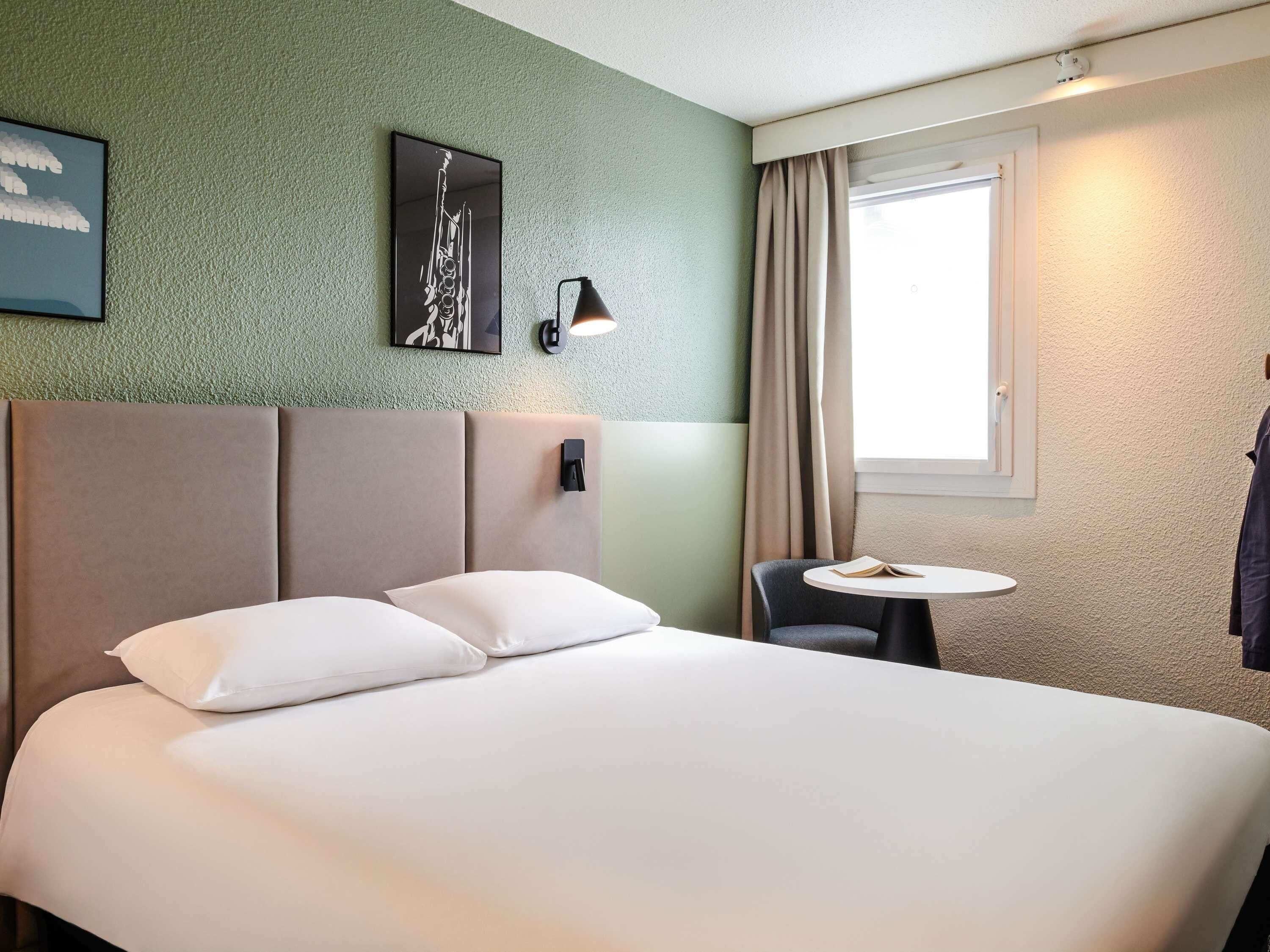 Une chambre d’hôtel avec un lit, une petite table, une chaise, une lampe fixée au mur et une fenêtre avec des rideaux.