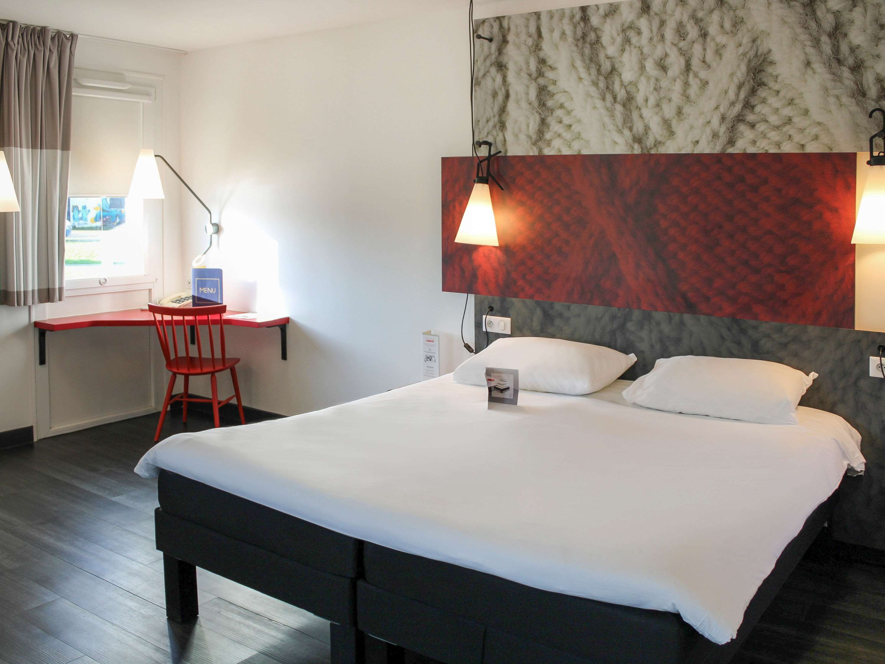 Une chambre d’hôtel avec un lit, un bureau et une chaise. Il y a une fenêtre avec des rideaux, un mur avec un motif texturé et deux lampes fixées au mur.