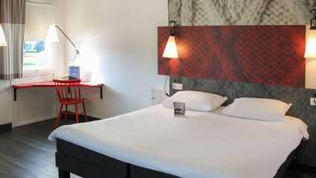 A hotel room with a bed, a desk, and a chair. There is a window with curtains, a wall with a textured pattern, and two wall-mounted lamps.