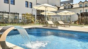 A swimming pool with a water feature, surrounded by lounge chairs and umbrellas, with a building in the background. - ibis La Roche sur Yon (Mouilleron-le-Captif)