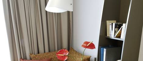 A hotel room with a laptop on a round table, a vase with a flower, a bookshelf, and a bed with pillows.