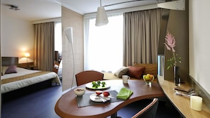 Een hotelkamer met een bed, een eettafel met ontbijtartikelen, een stoel, een bureau met een lamp en een vaas met bloemen.