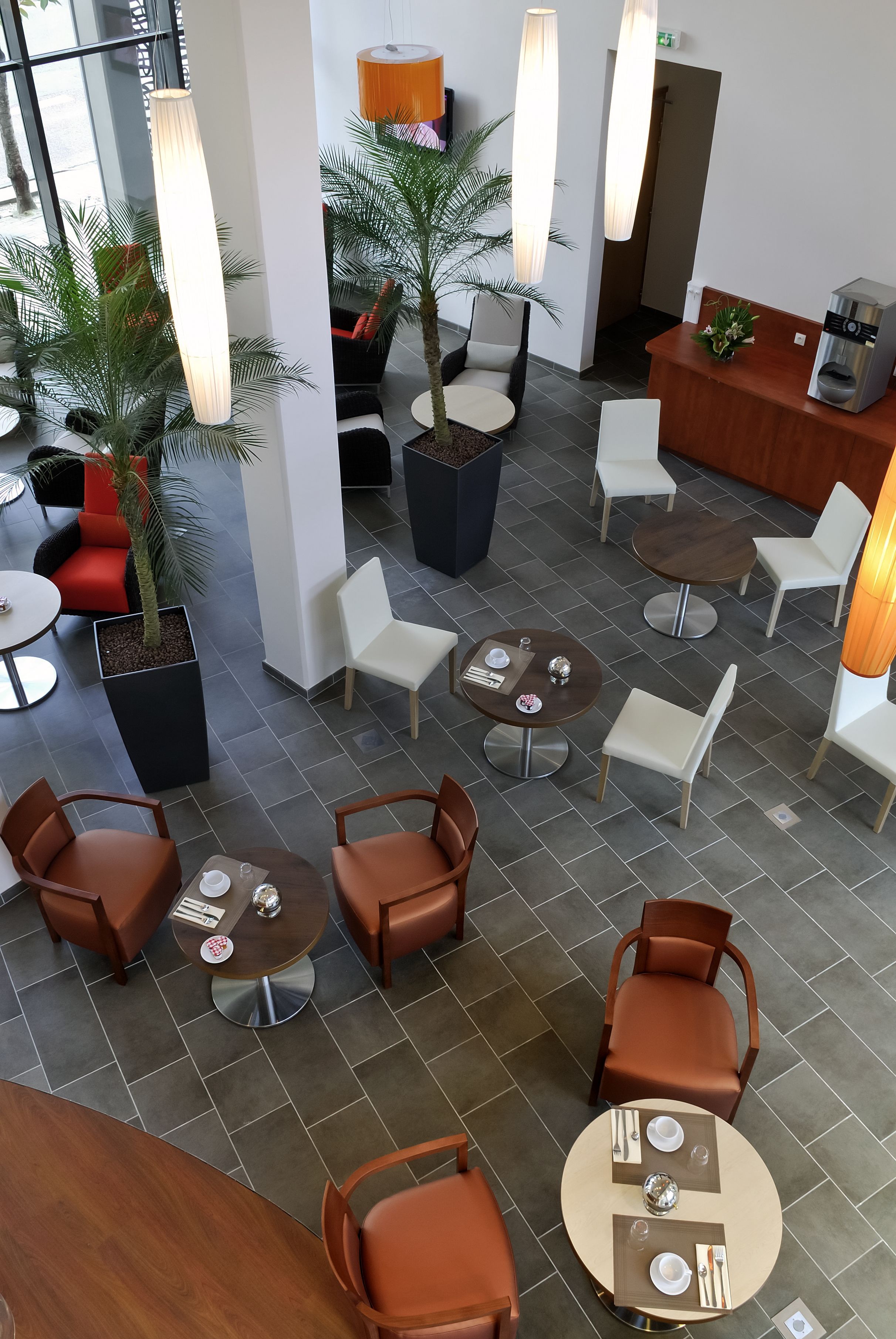 A modern hotel lobby with multiple seating areas, including round tables and chairs, and a reception desk with a coffee machine.