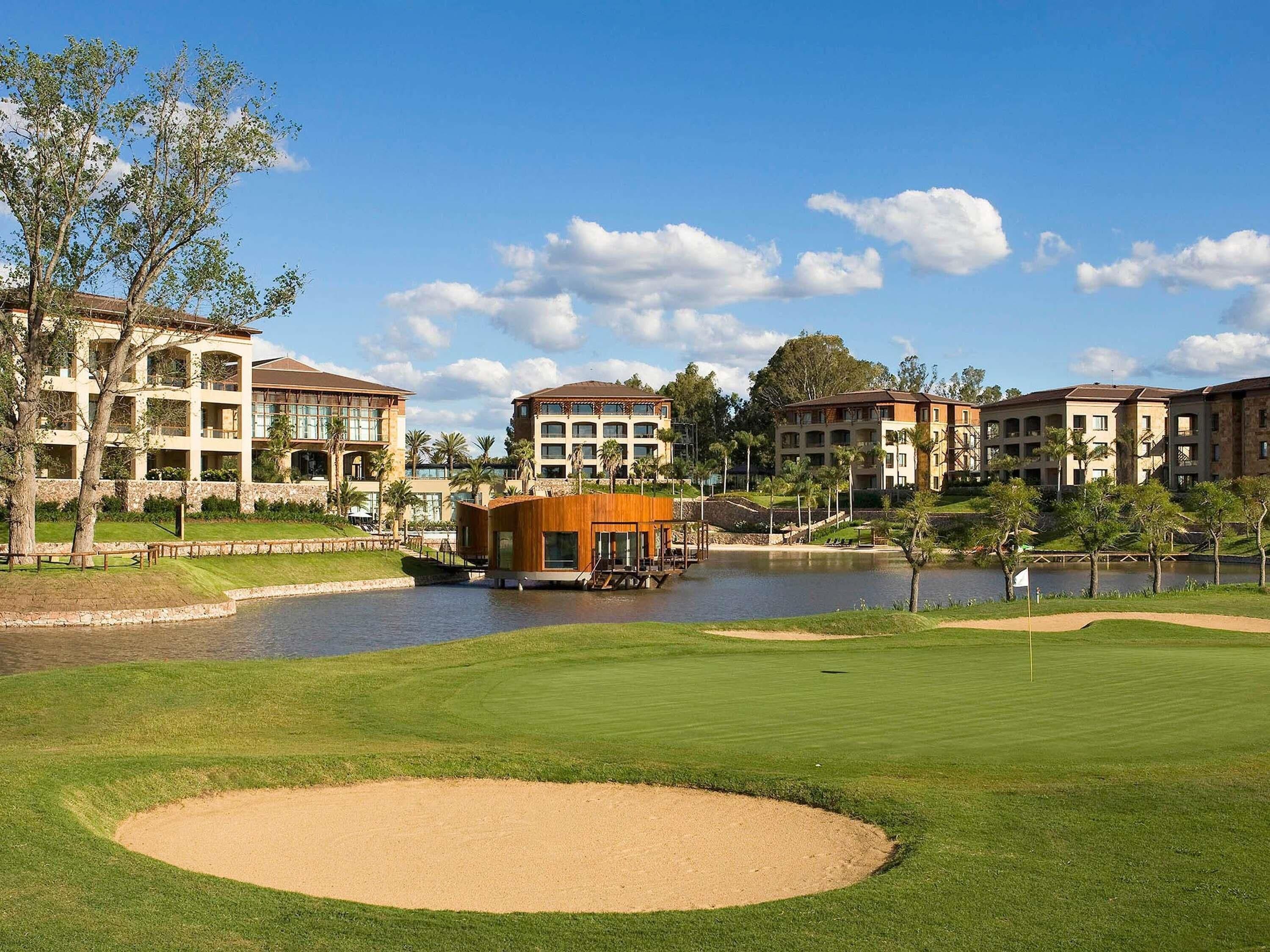 A golf course with a sand bunker, a pond, and multi-story buildings in the background.