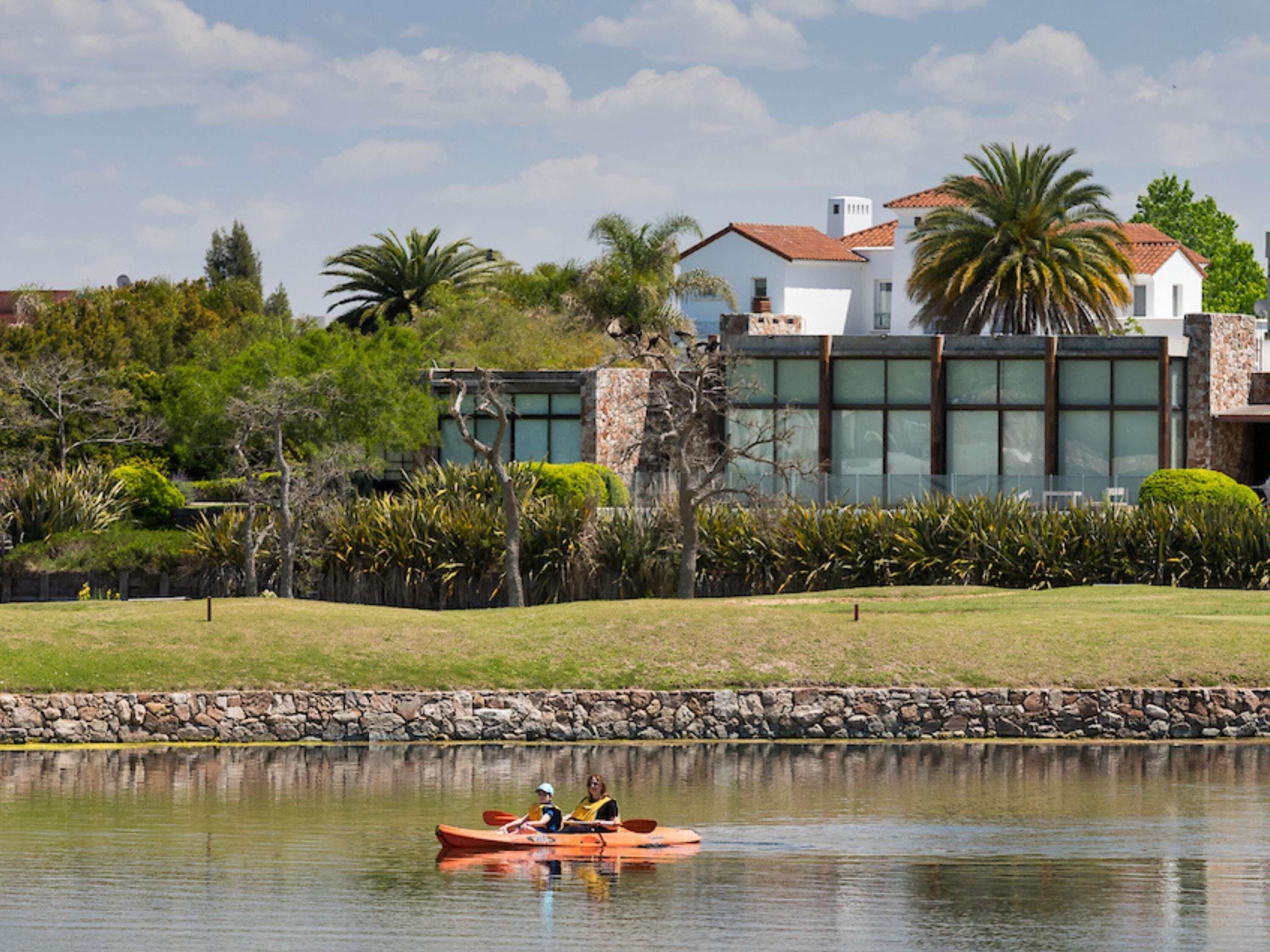 A calm lake with a kayak, a stone wall, and a modern building with large windows.