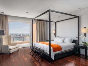 A four-poster bed with a white and orange bedspread, a chair, a nightstand, and a view of the city through large windows. - Sofitel La Reserva Cardales (Campana)