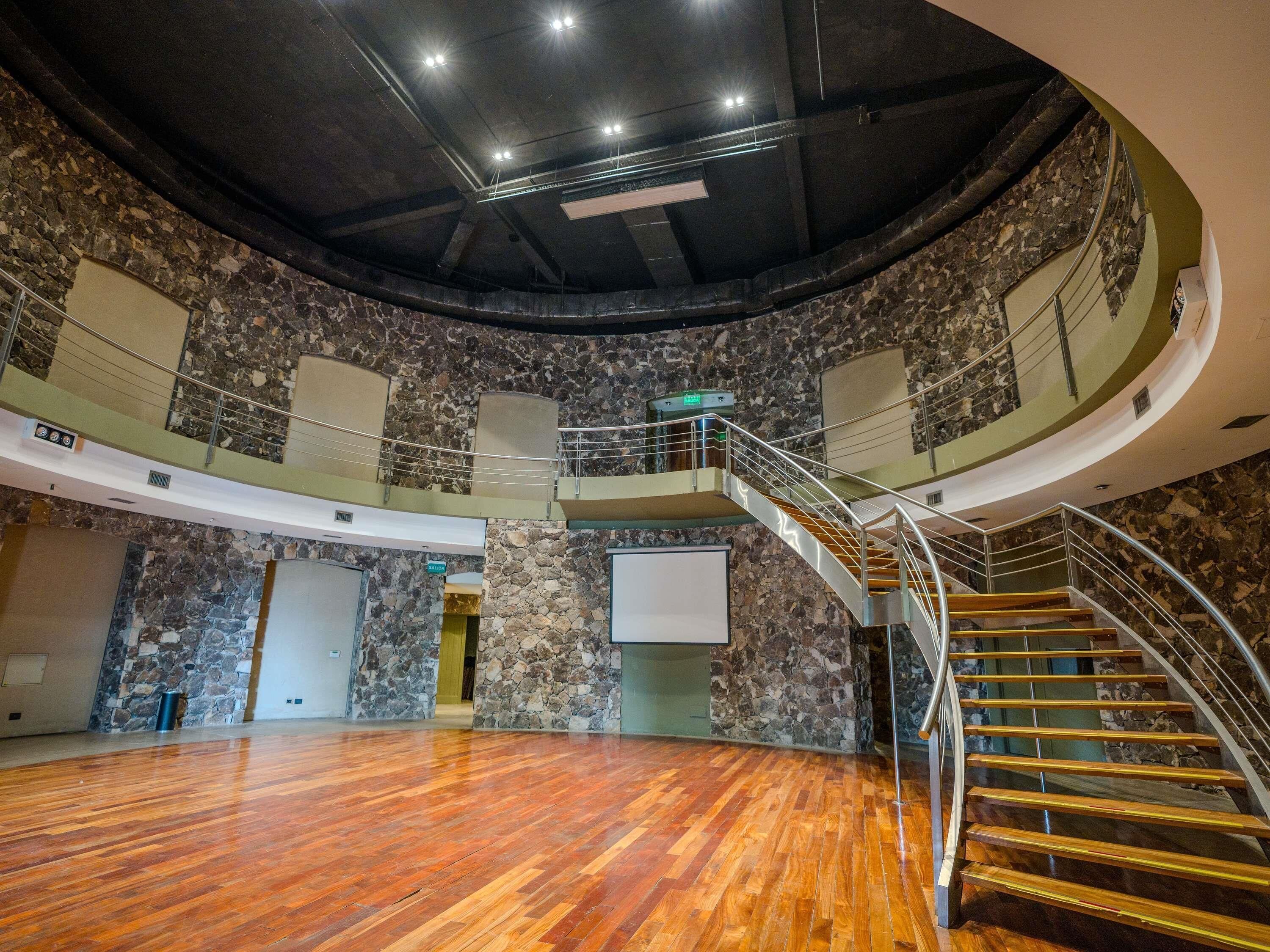 A spacious hall with a wooden floor, a curved staircase, and stone walls.